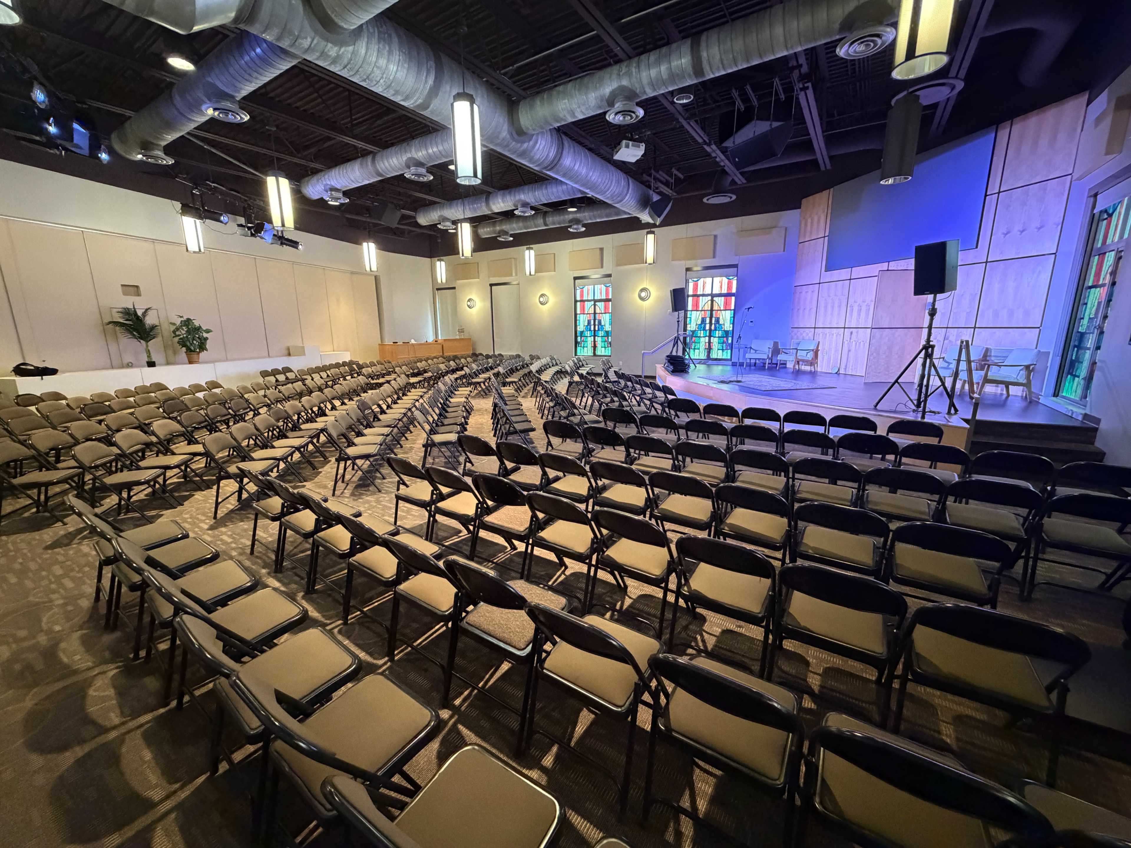 The image shows a spacious auditorium with rows of black folding chairs arranged in front of a small stage equipped with a microphone and colorful stained-glass windows.