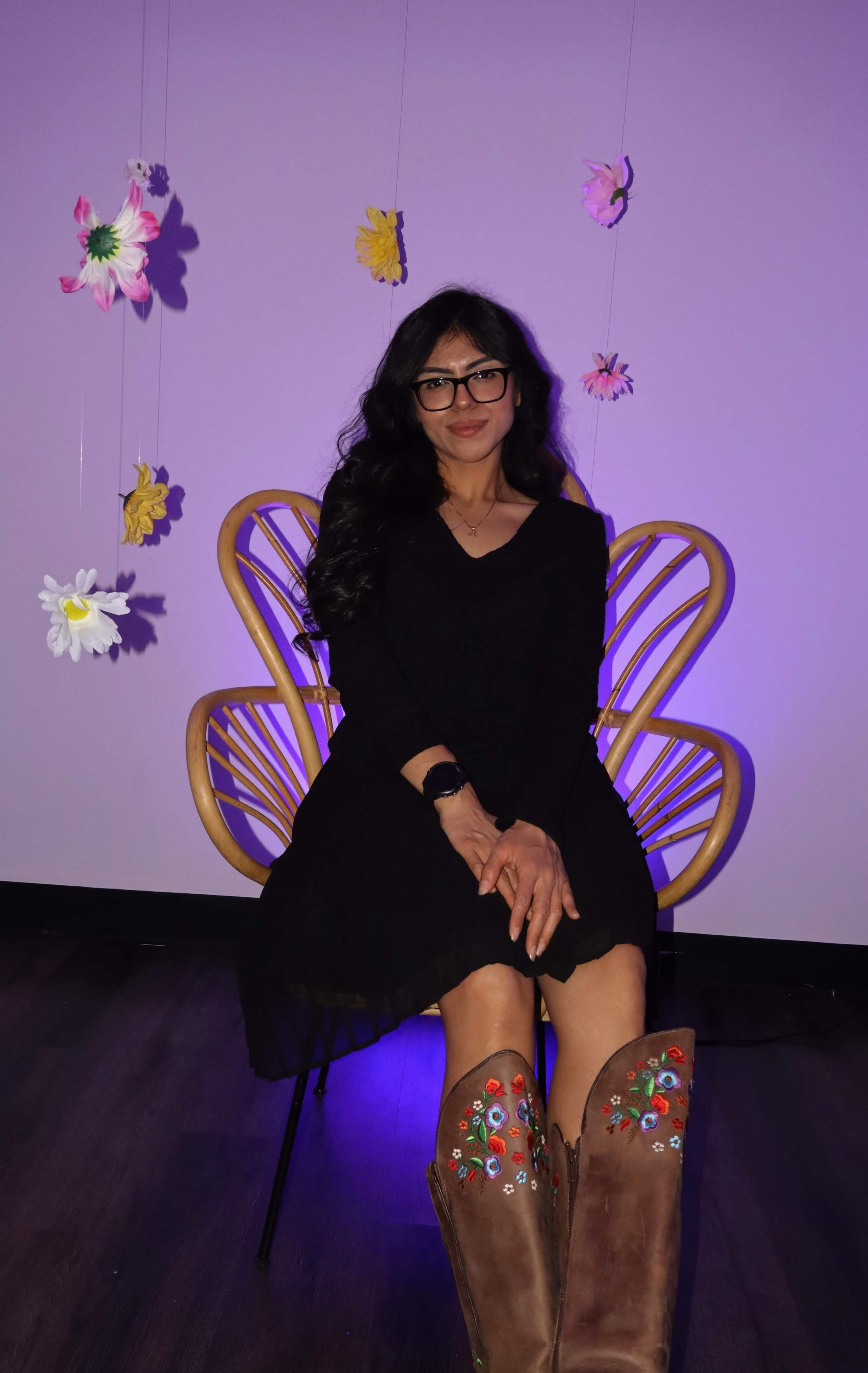A woman with long black hair sits on a chair adorned with flowers hanging from the ceiling, wearing a black dress and embellished brown boots.