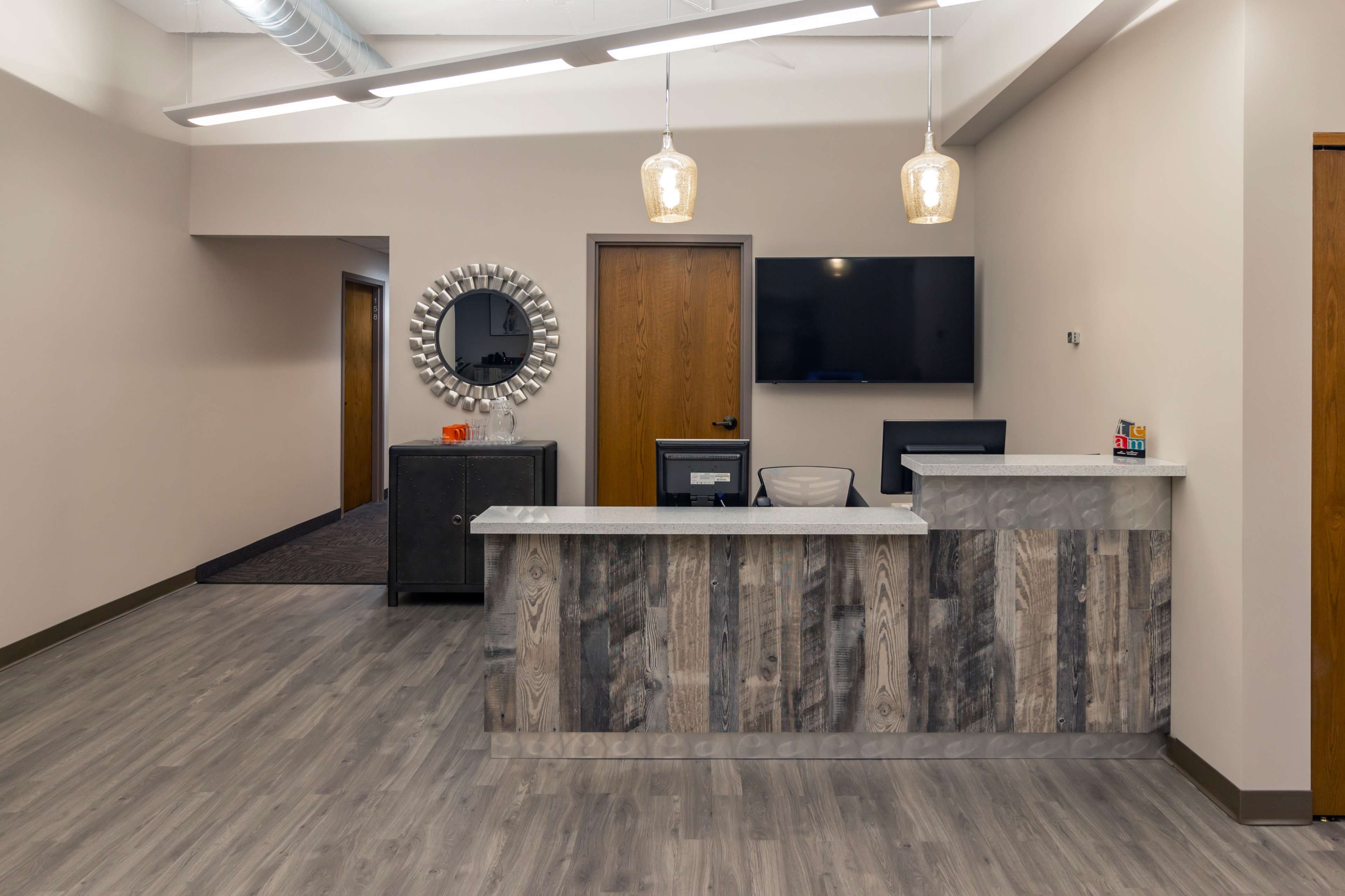 A modern reception area featuring a wooden front desk, a large round mirror, and a wall-mounted TV in a well-lit space.