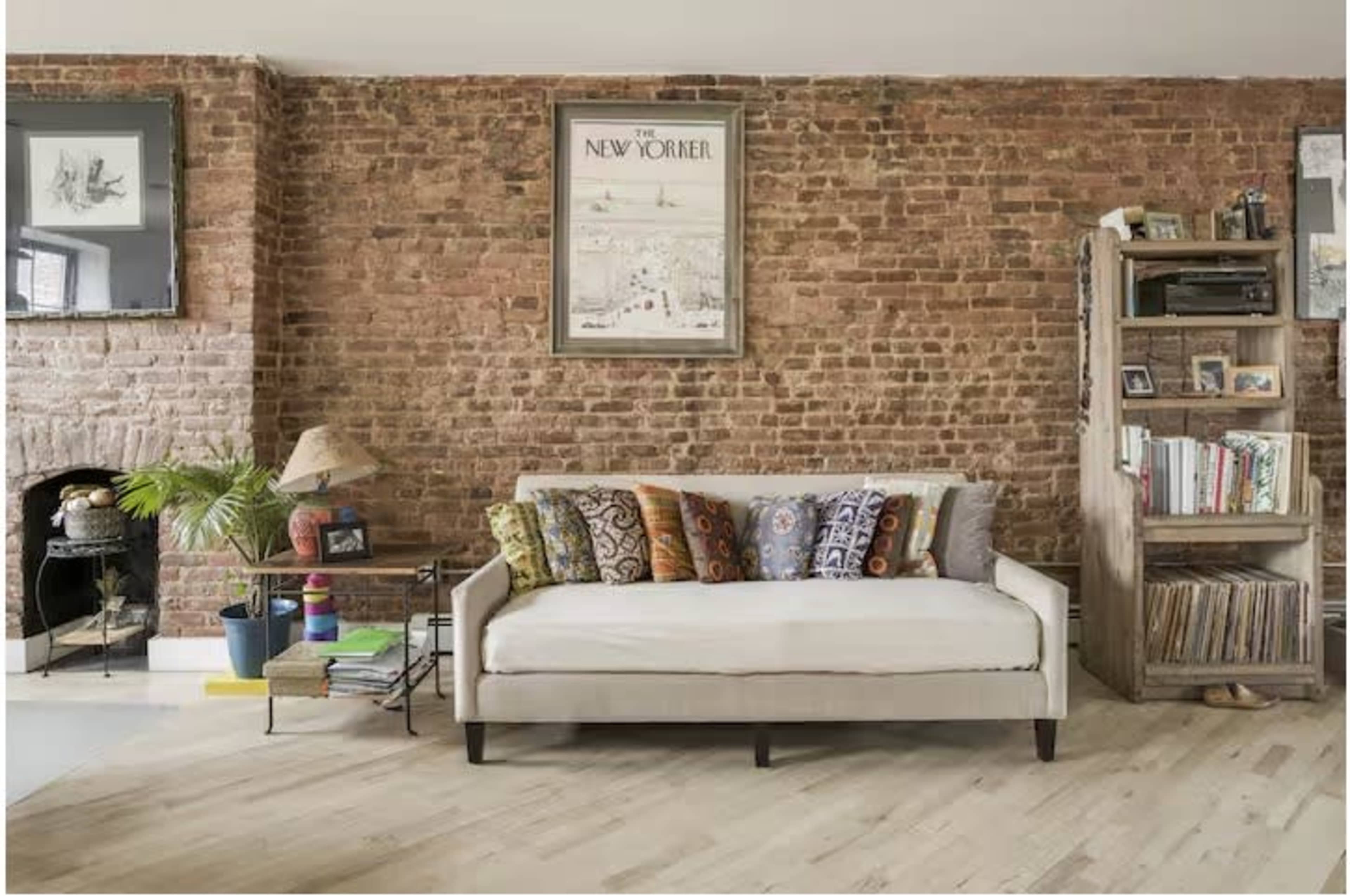 A beige sofa with colorful pillows sits against a textured brick wall, beside a wooden bookshelf filled with books and decorations.