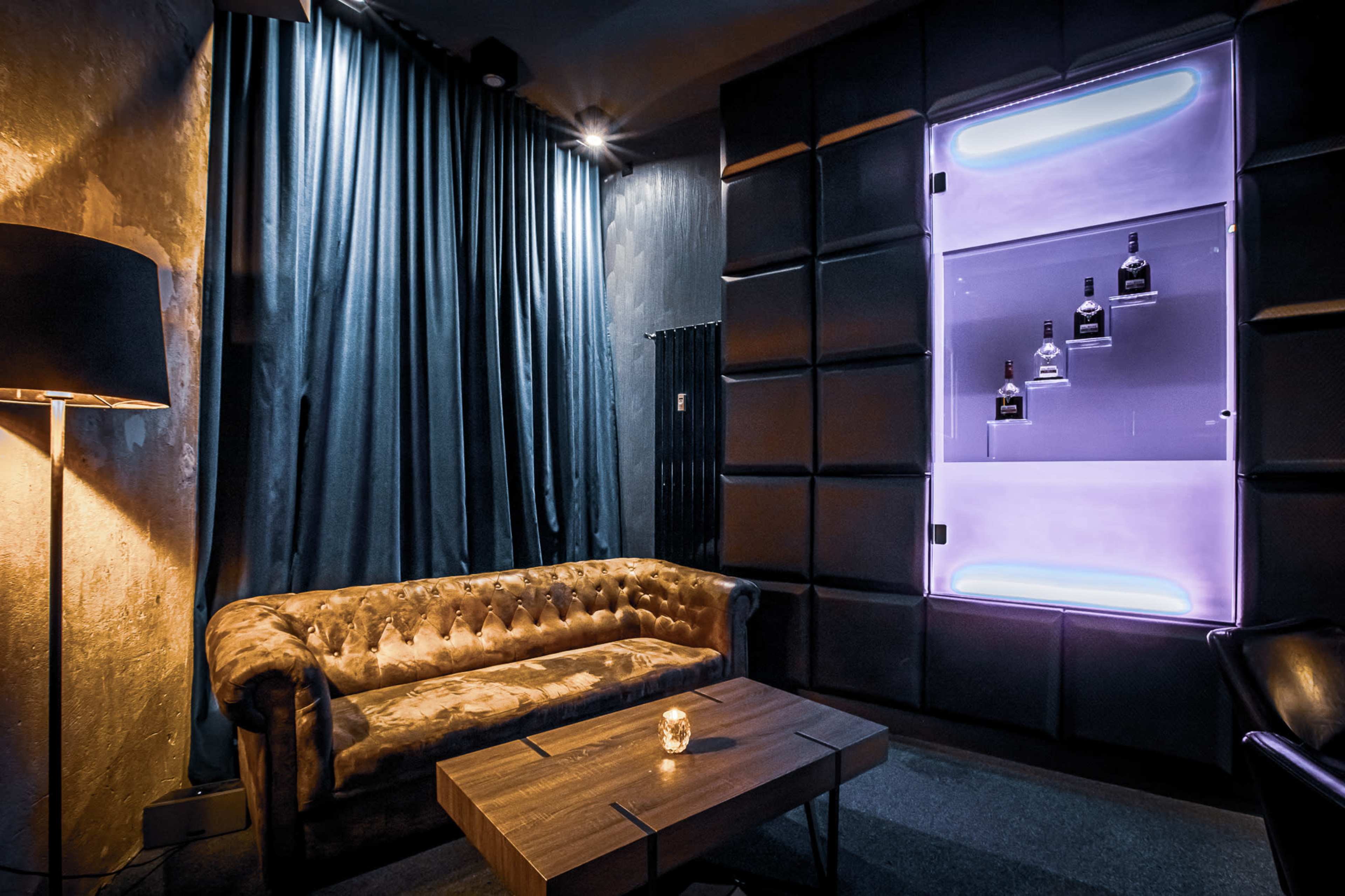 The image shows a dimly lit lounge area with a tufted brown sofa, a wooden coffee table, and a wall display featuring illuminated bottles.