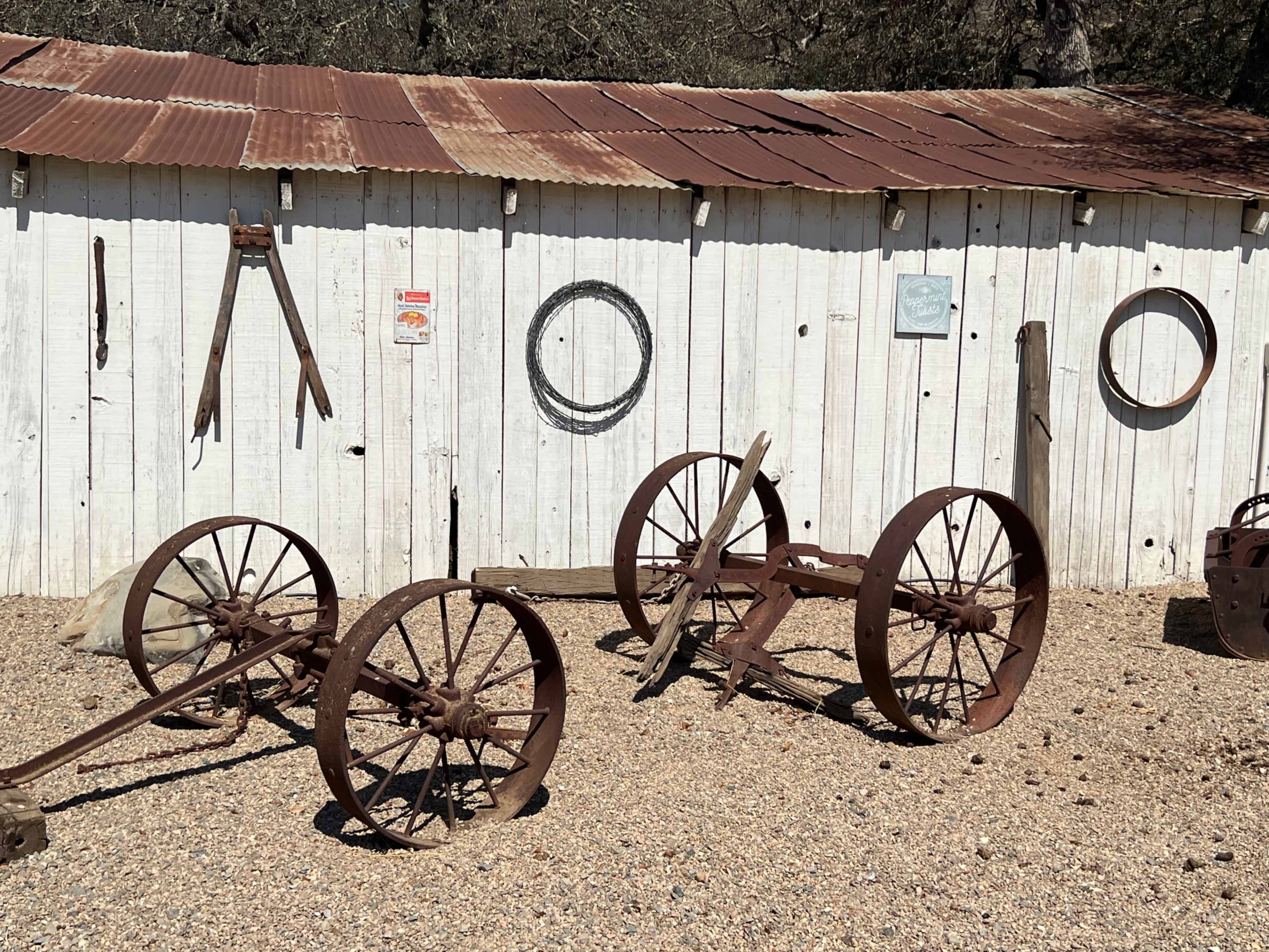 An assortment of vintage farm equipment and tools is displayed against a rustic, whitewashed shed with a corrugated metal roof.