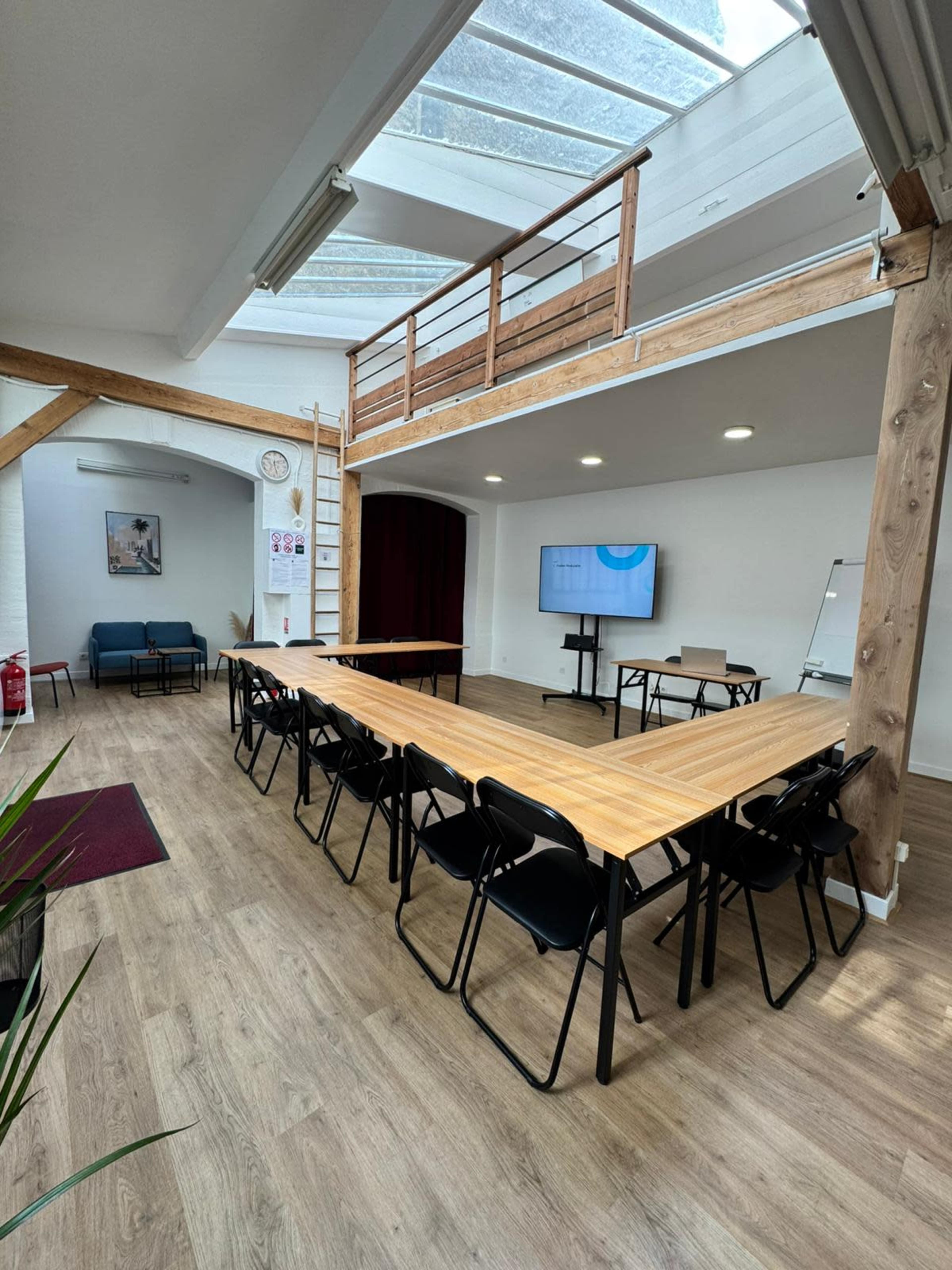 The image shows a spacious, well-lit meeting room with a large wooden table arranged in a V-shape, surrounded by chairs, and featuring a screen on one wall.
