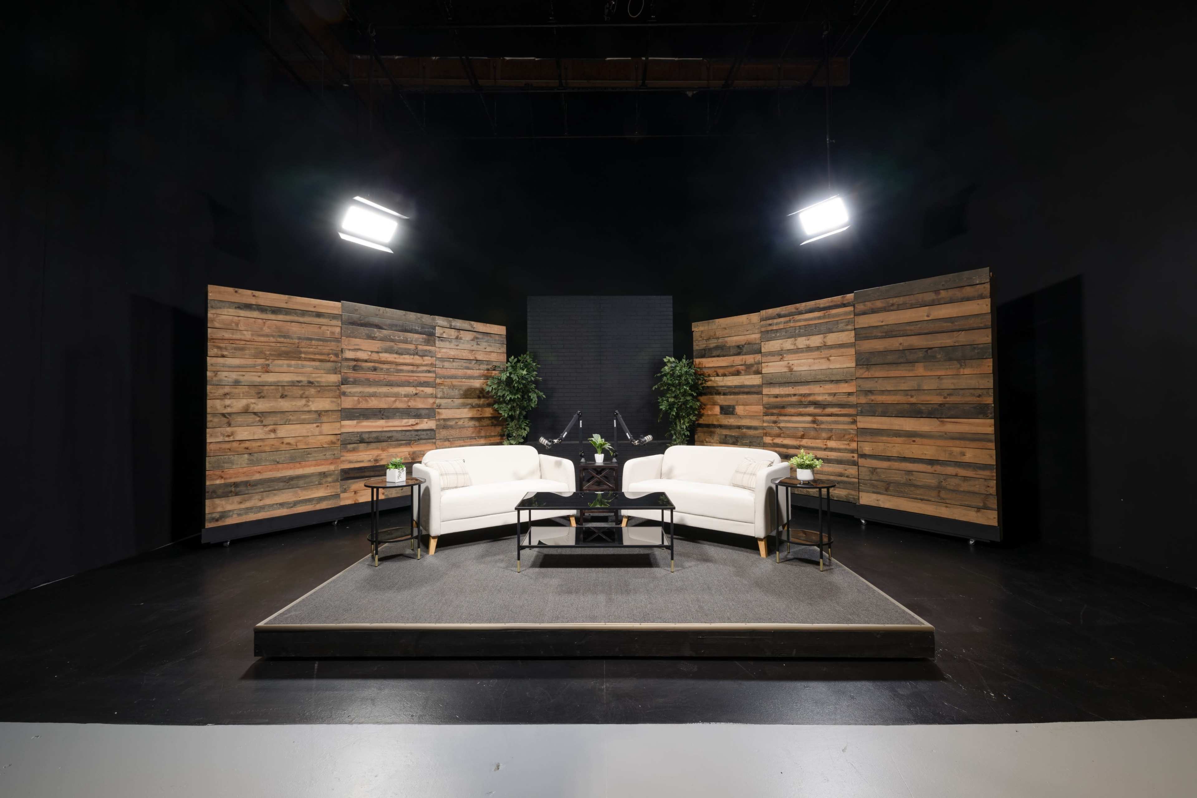 A modern media studio setup featuring two white sofas, a black coffee table, and wooden partition walls, with large lights positioned above.