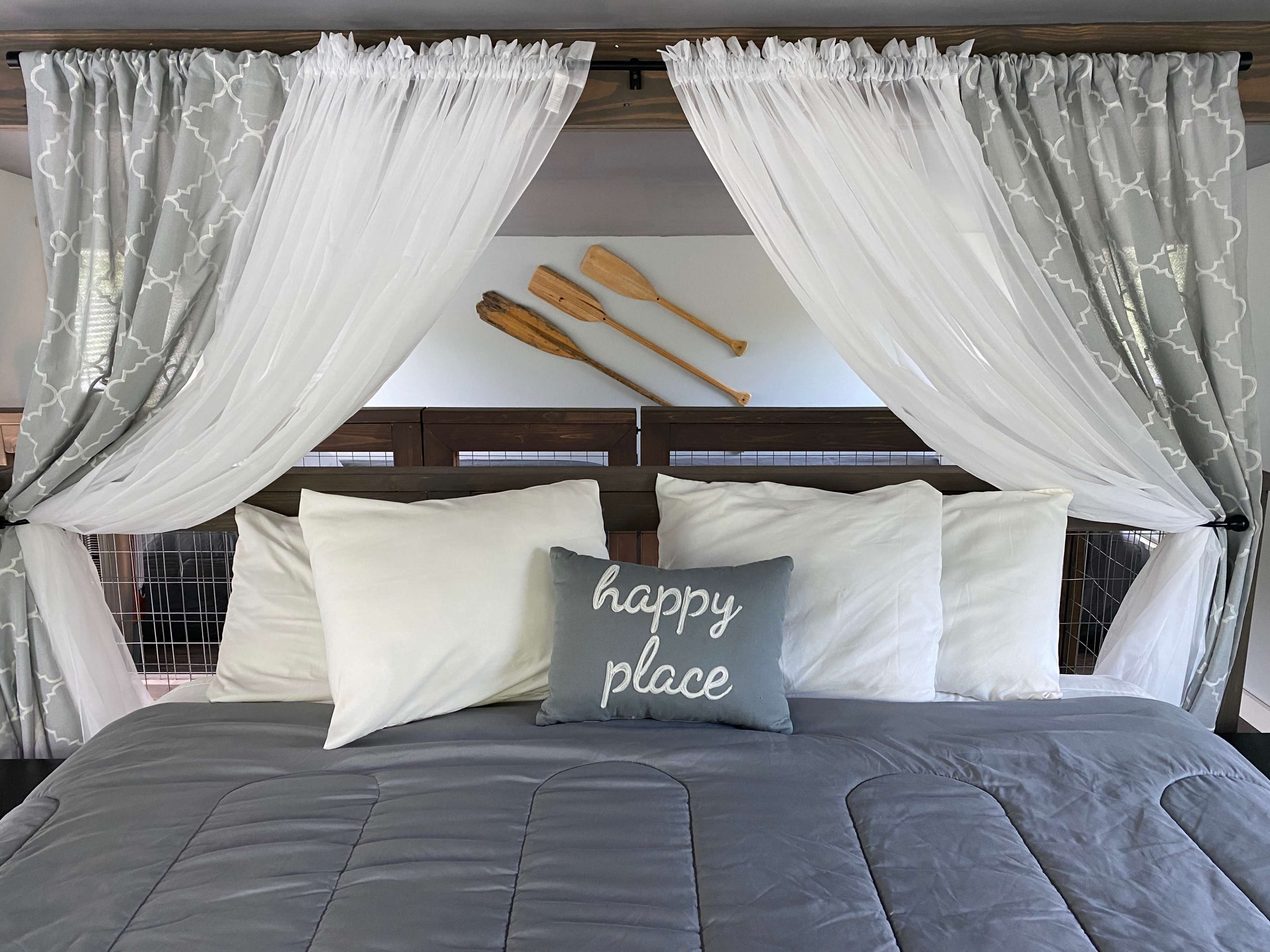 A neatly arranged bed with gray bedding, multiple white pillows, and sheer curtains, adorned with decorative oars on the wall above.