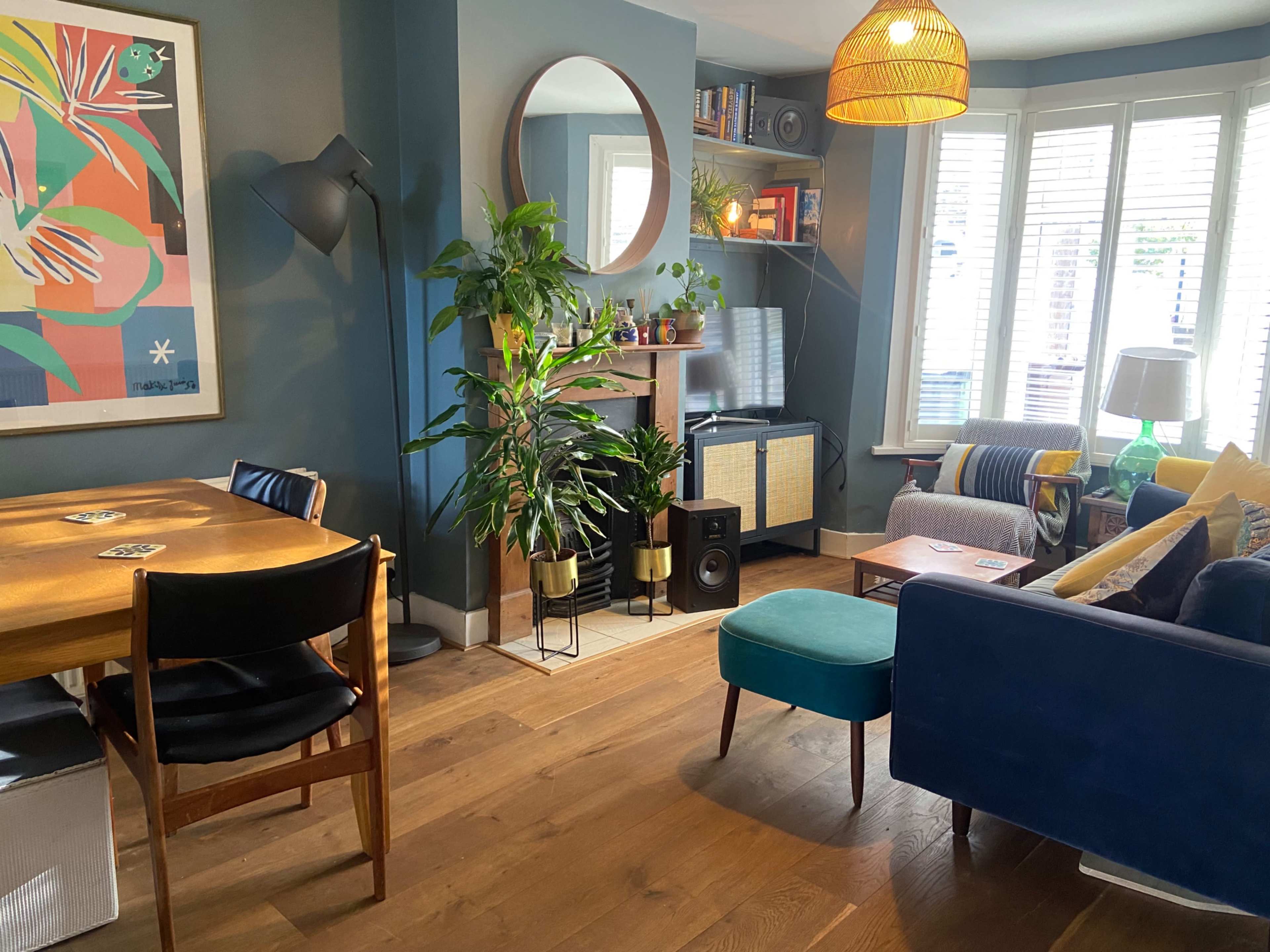 A cozy living room with a wooden dining table, a plush sofa, a large mirror, and decorative plants, all arranged around a bright window.