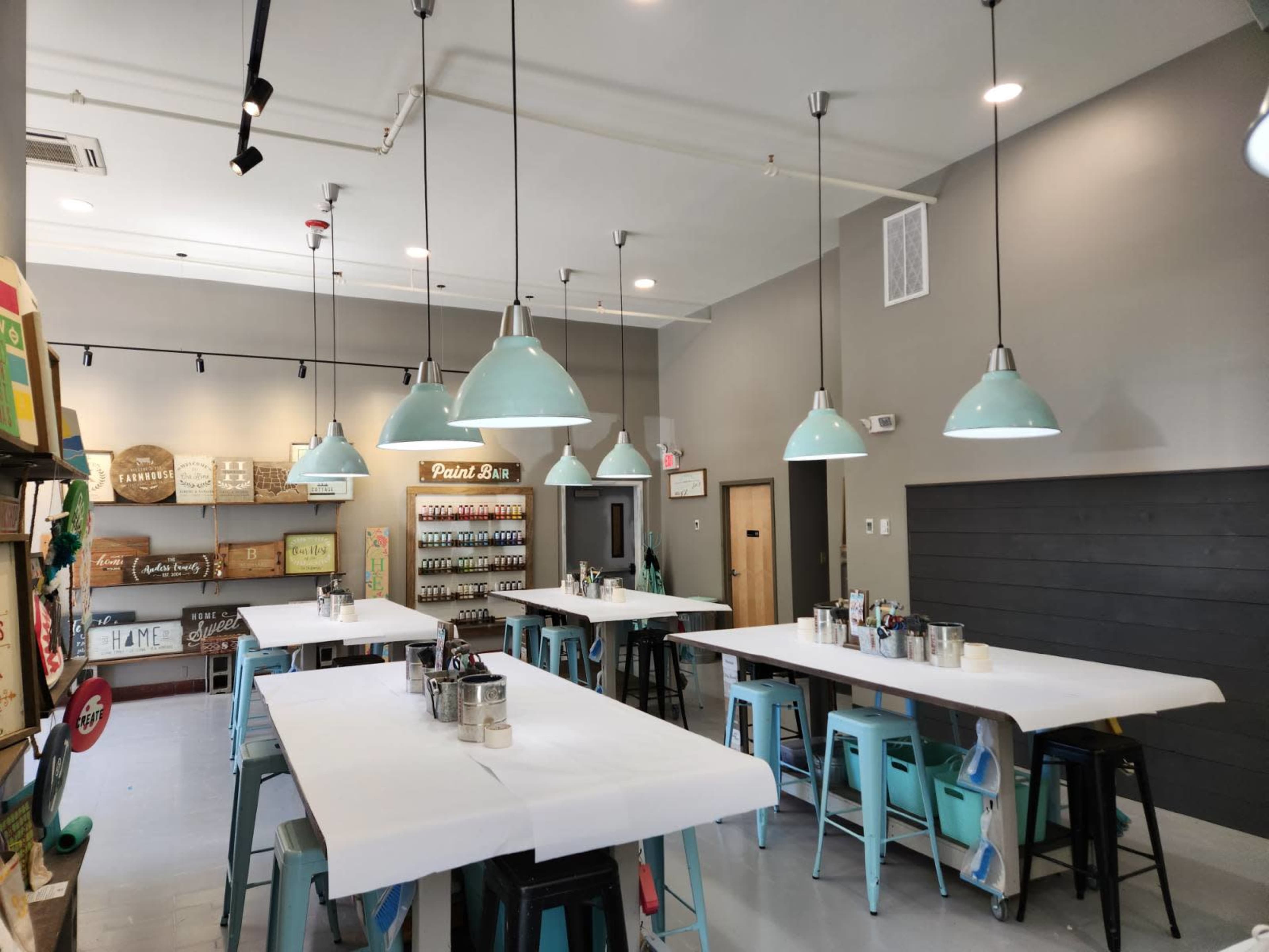 A brightly lit art studio with several tables covered in white paper, surrounded by blue pendant lights and shelves displaying paint supplies.