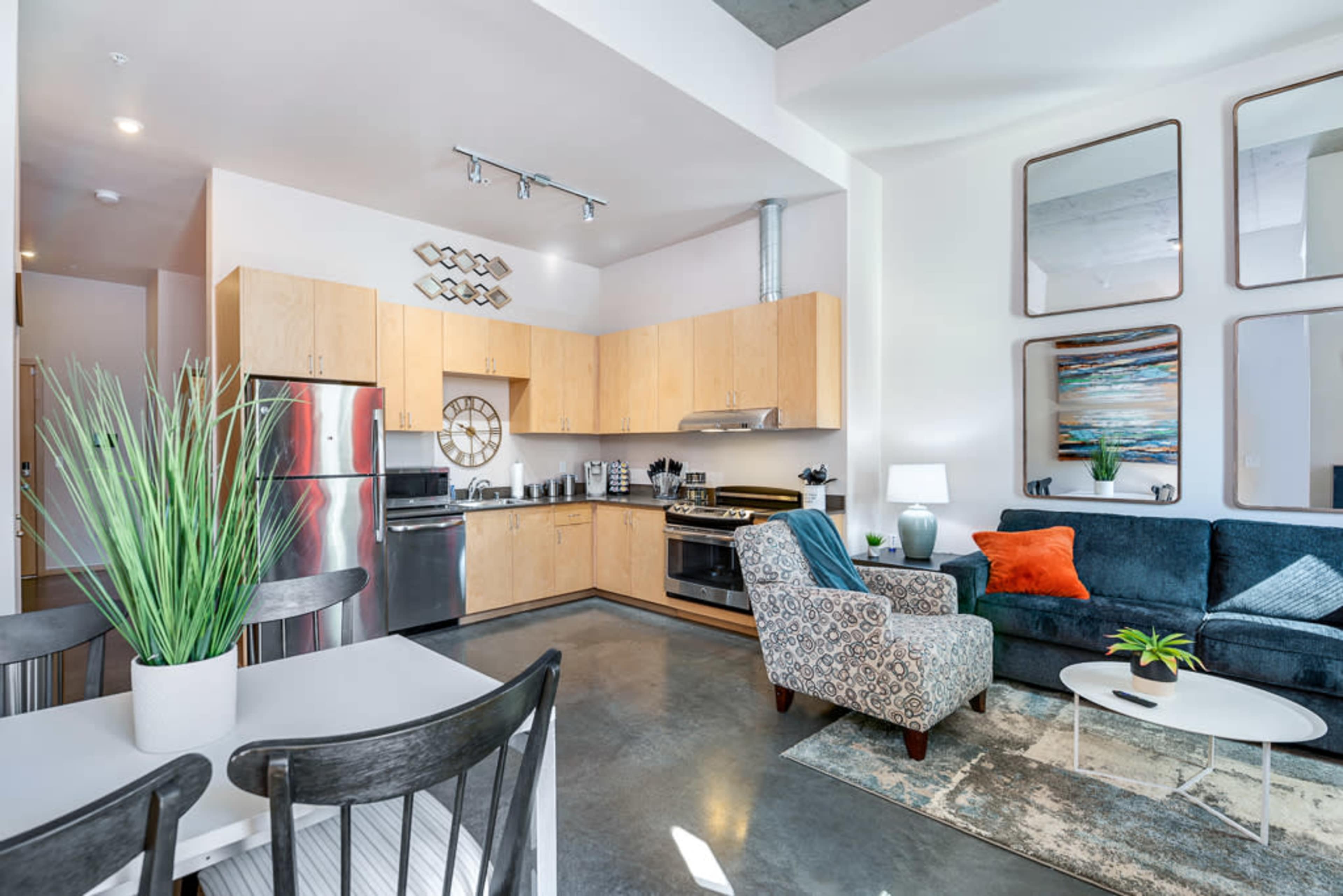 The image shows a modern kitchen and living area featuring wooden cabinets, stainless steel appliances, a blue sofa, and a dining table with black chairs.