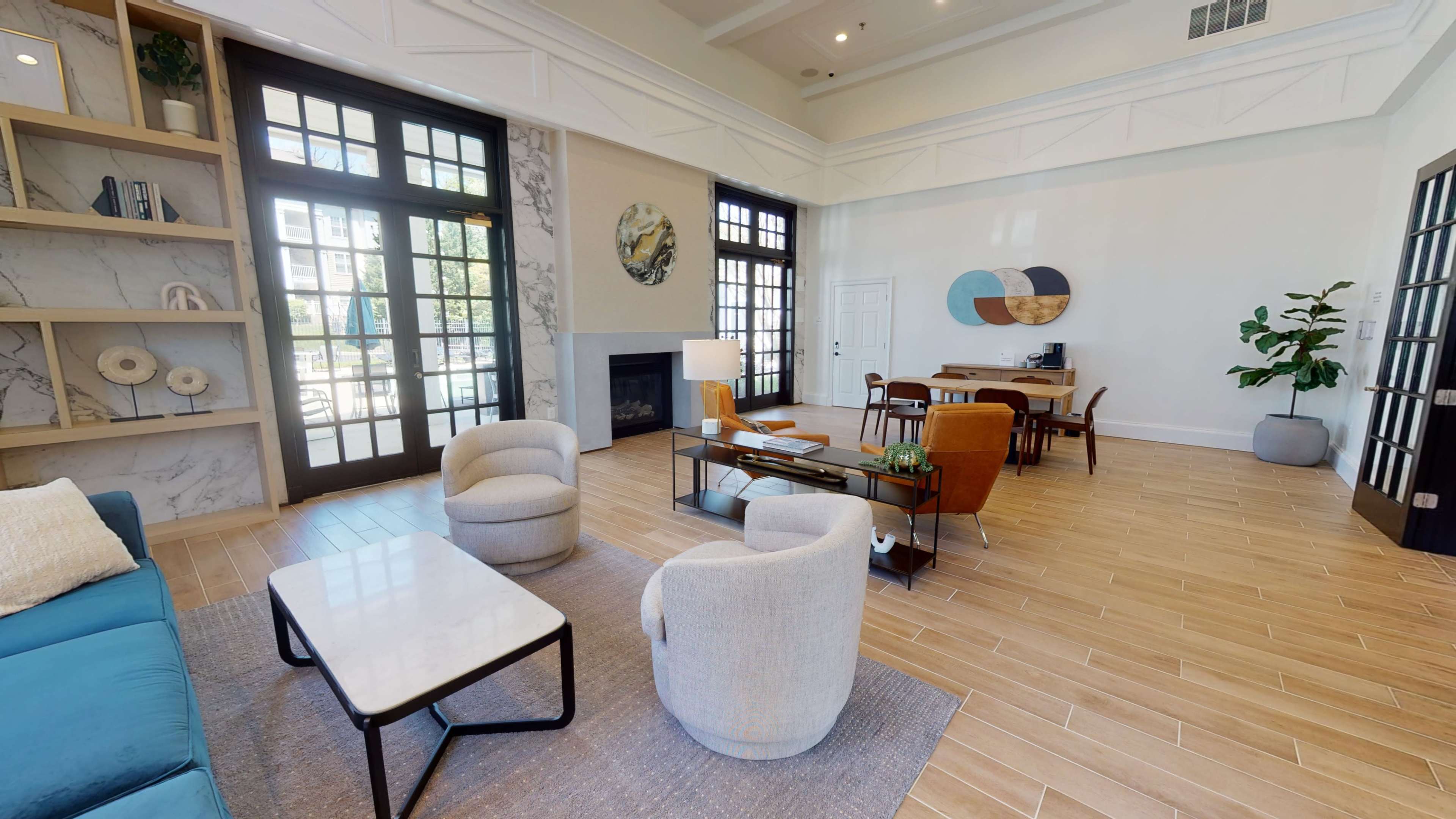 The image shows a spacious, bright common area featuring a seating arrangement with two armchairs and a sofa, a coffee table, a fireplace, and dining space with a table and chairs.