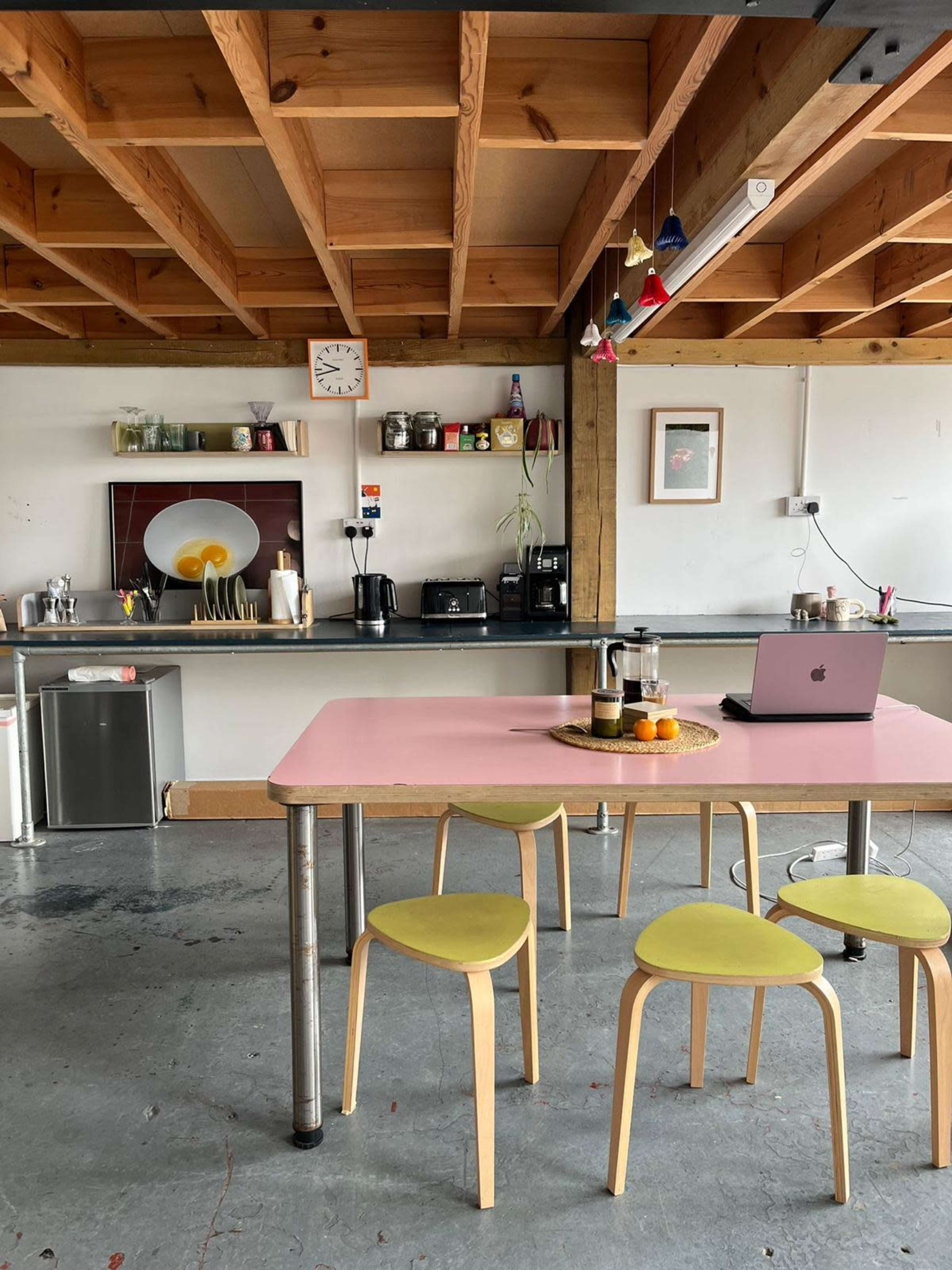 The image shows a modern kitchen with a pink table and yellow chairs, featuring a coffee maker, a clock, and various kitchenware on the shelves.