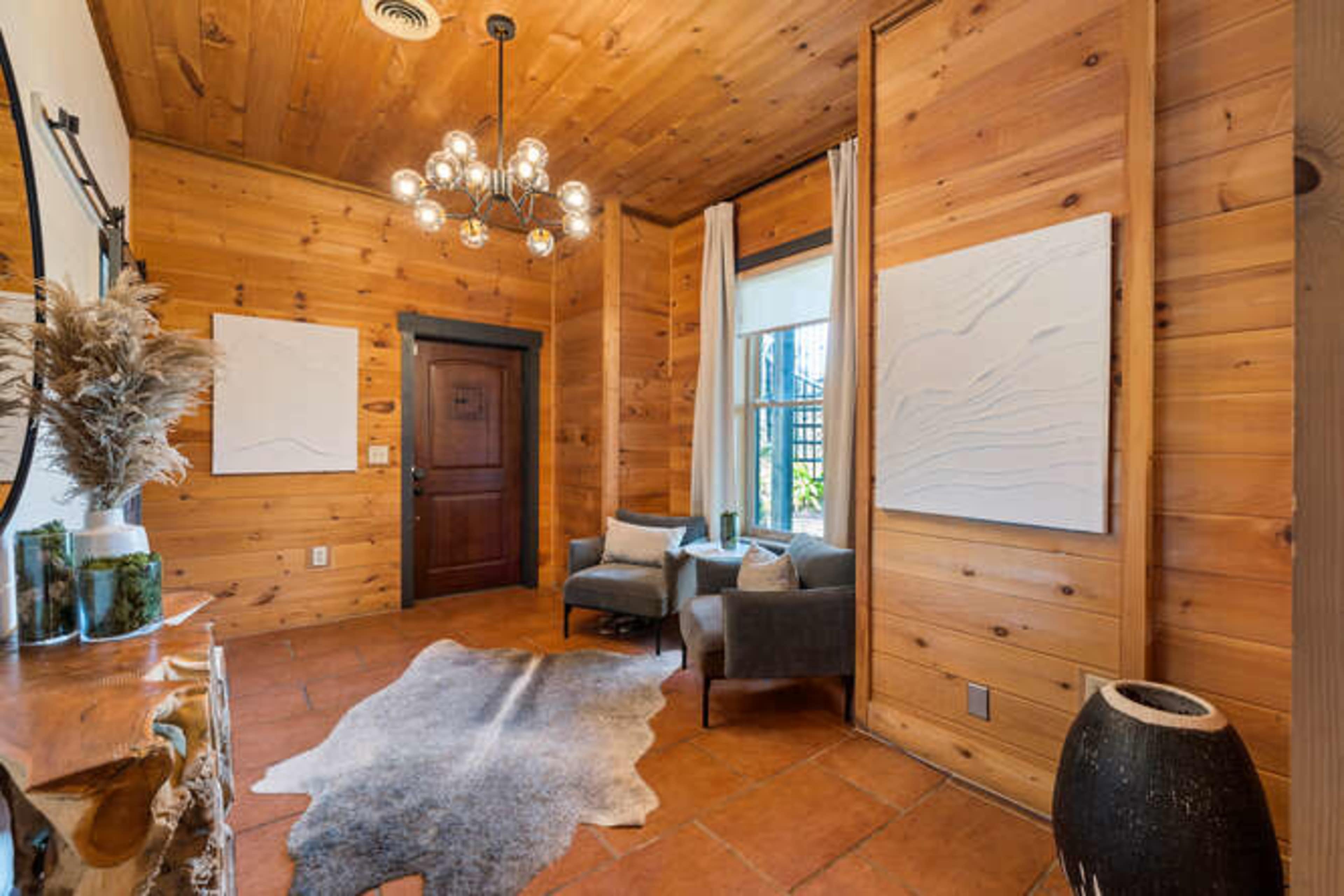 A corner of a wooden-paneled room featuring two gray chairs, a decorative rug on the tiled floor, and abstract wall art.