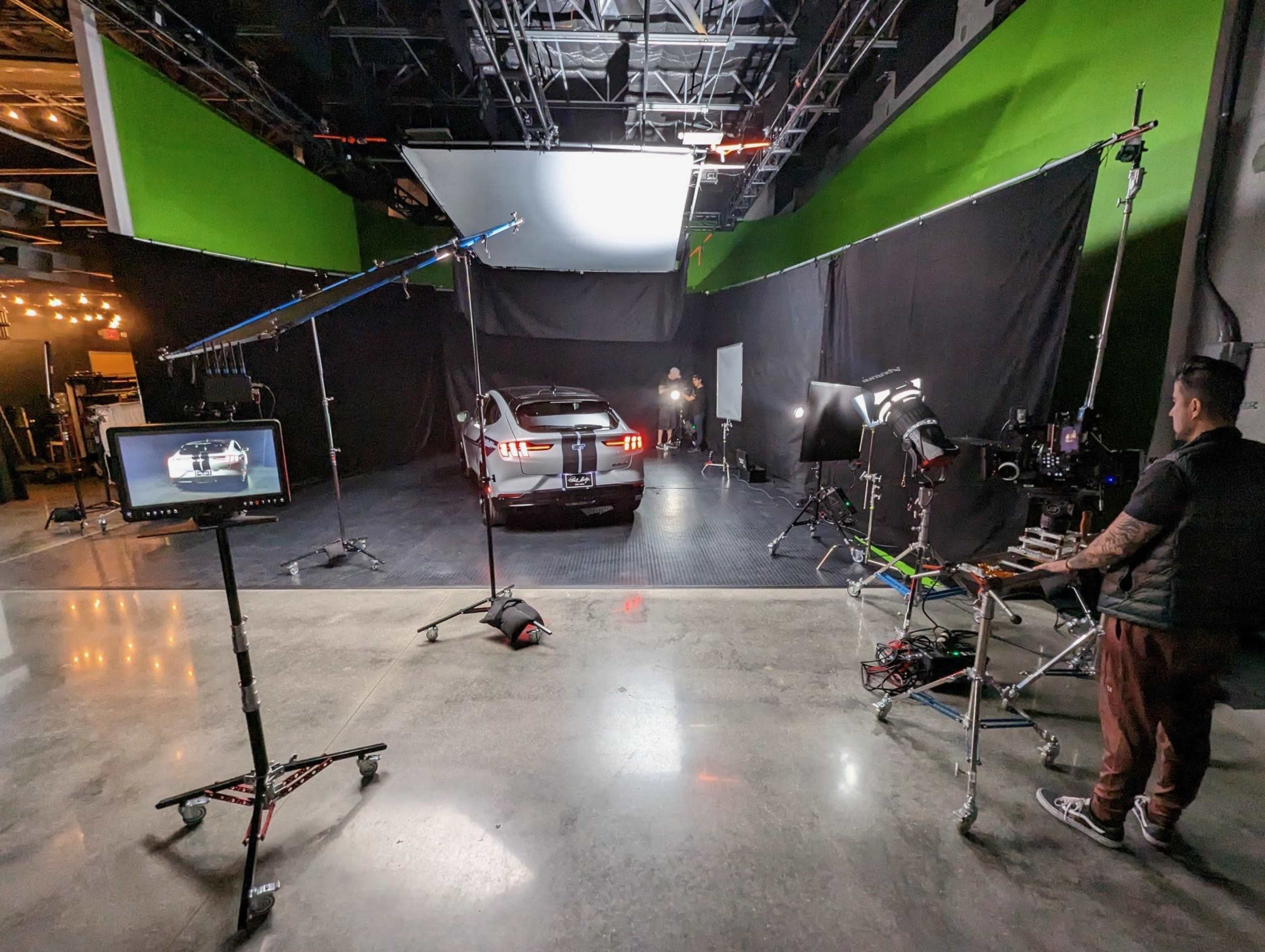 A film crew sets up lighting and equipment around a car in a studio with green screens and dark backgrounds.
