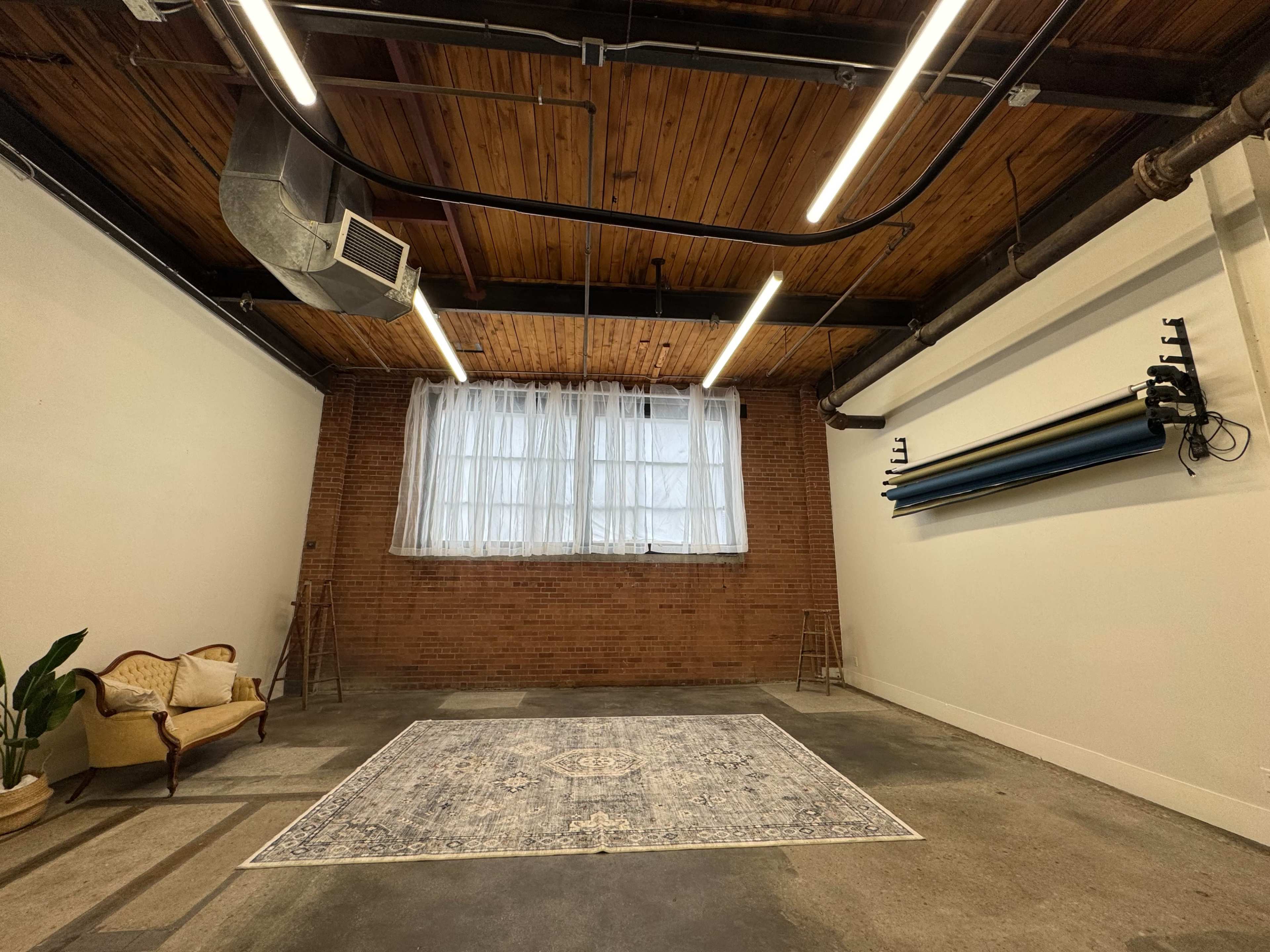 The image shows a spacious interior with exposed brick walls, wooden beams, and a large window, featuring a patterned rug centered on a concrete floor.