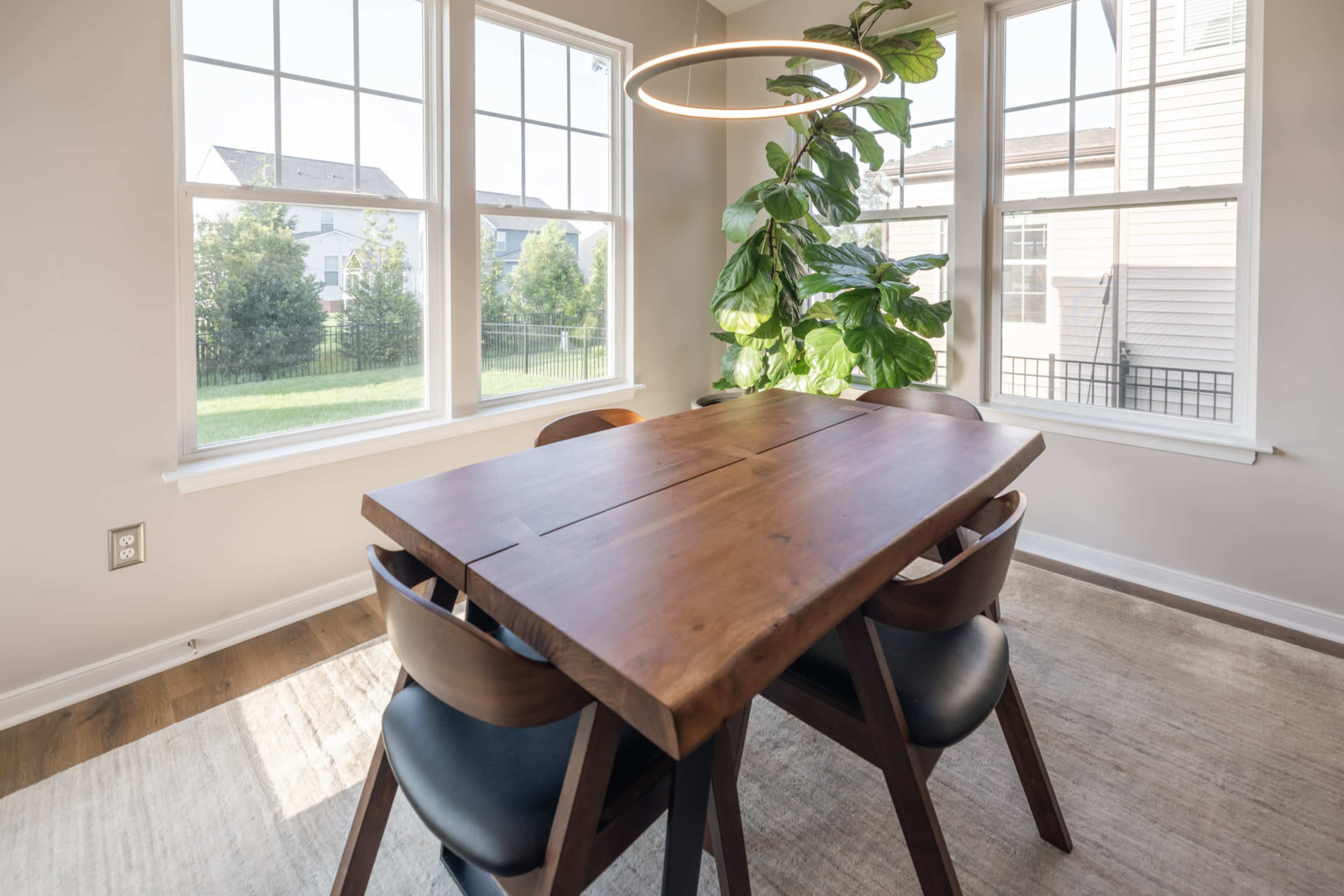 A wooden dining table with four chairs is positioned near large windows, with a hanging light fixture and a potted plant in the corner.