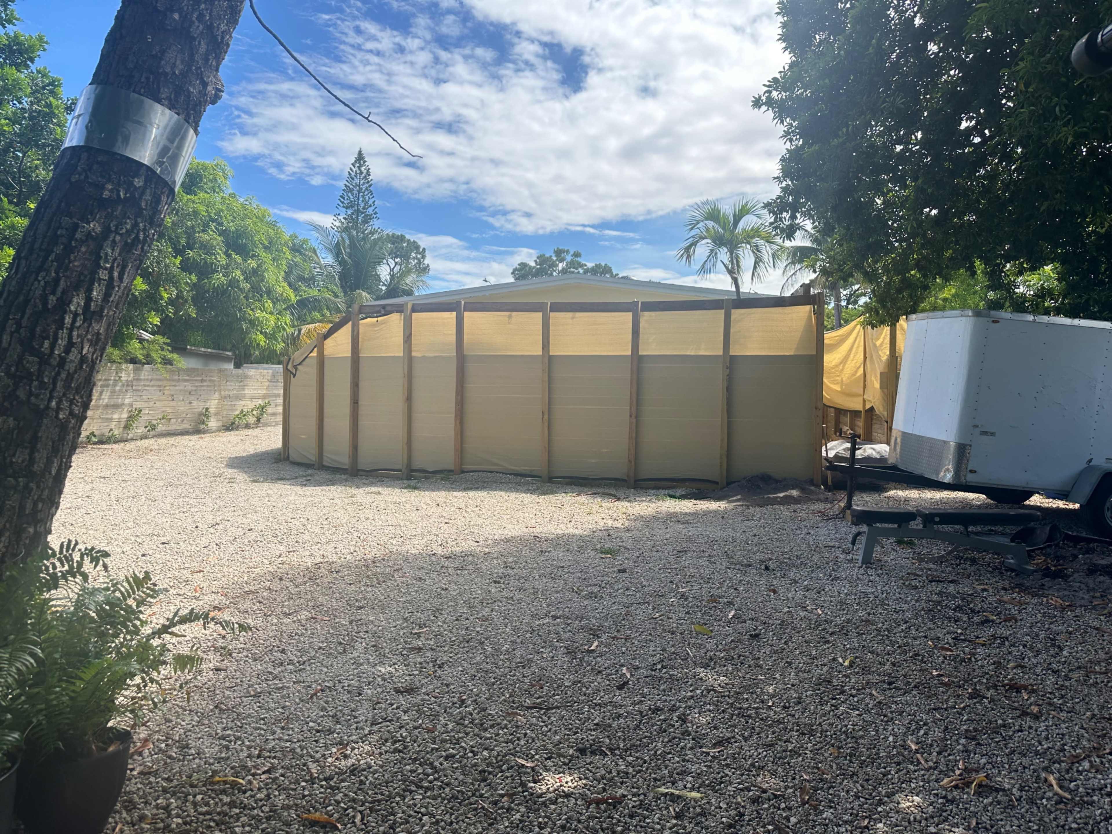 A circular structure with wooden framing and yellow tarp covers is set in a gravel area surrounded by trees and a trailer.