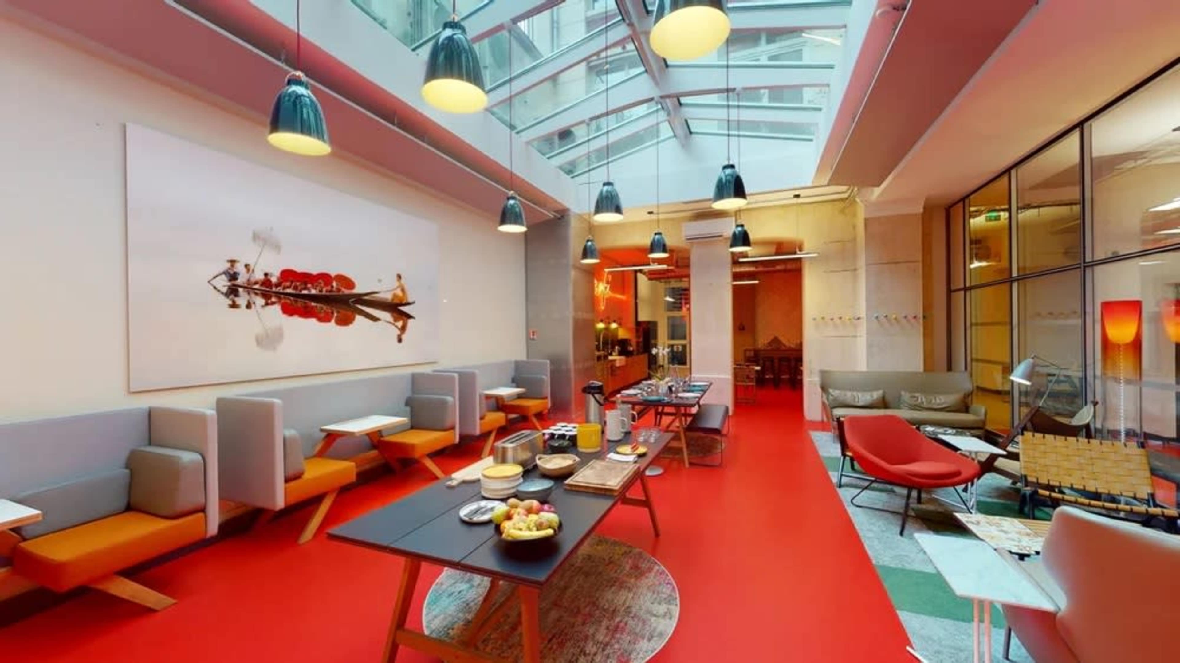 A modern open space with red flooring, large windows, and seating areas featuring gray and orange furniture along with a dining setup.