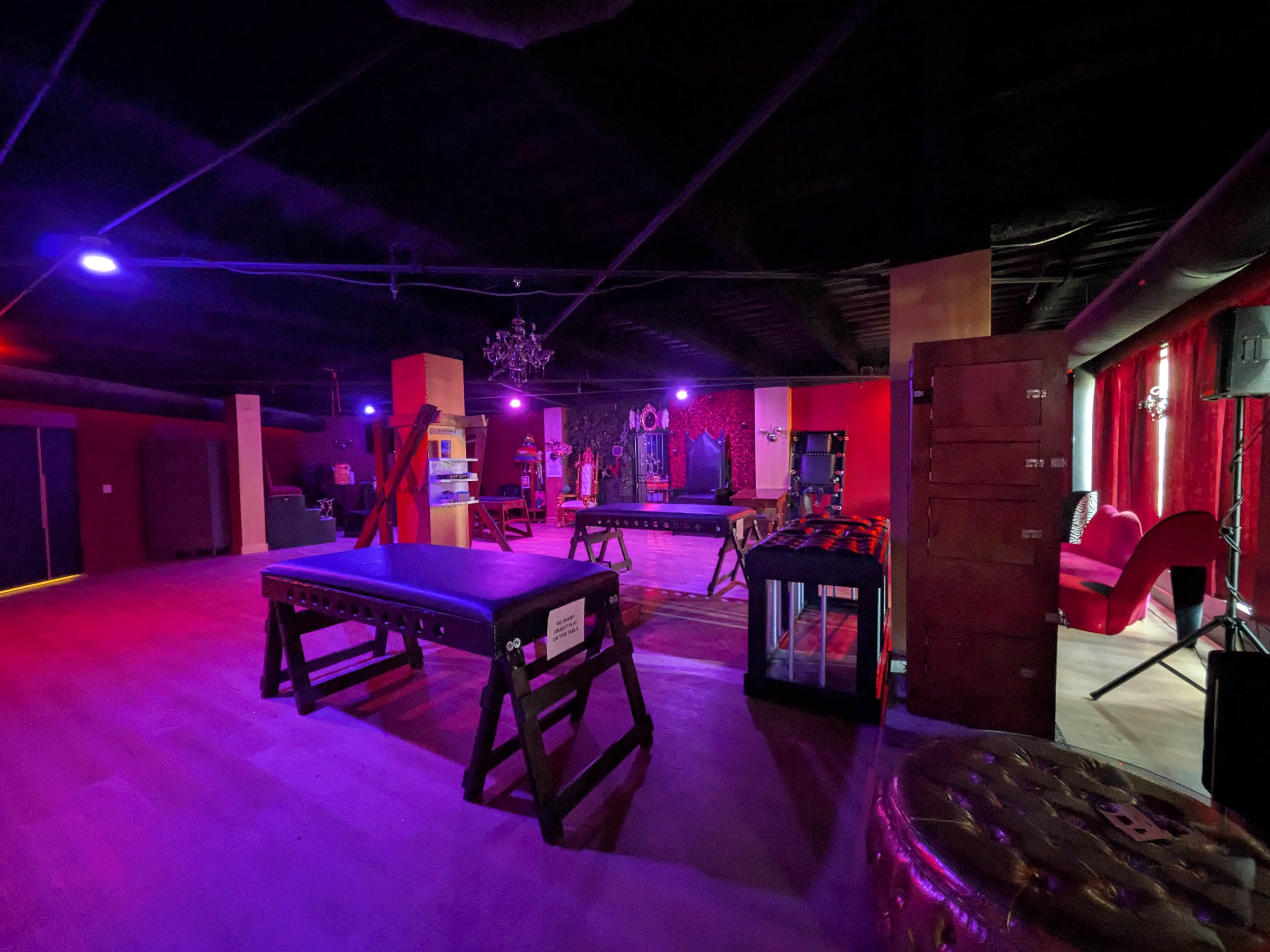 The image shows a dimly lit room with various pieces of equipment, including two massage tables, surrounded by red and purple lighting and modern furnishings.
