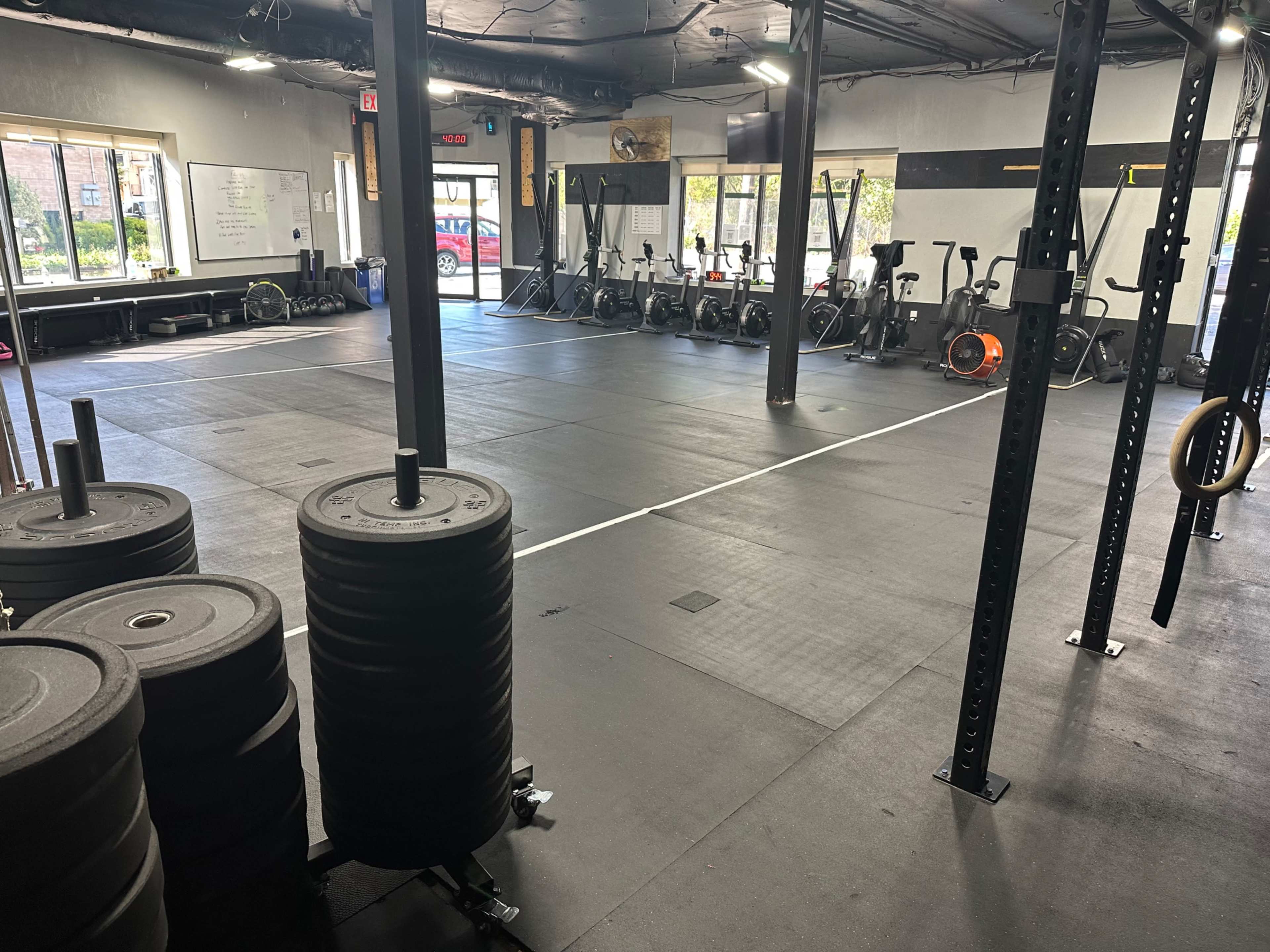 The image shows a gym with black rubber flooring, various fitness equipment, and weight plates stacked near a set of squat racks.