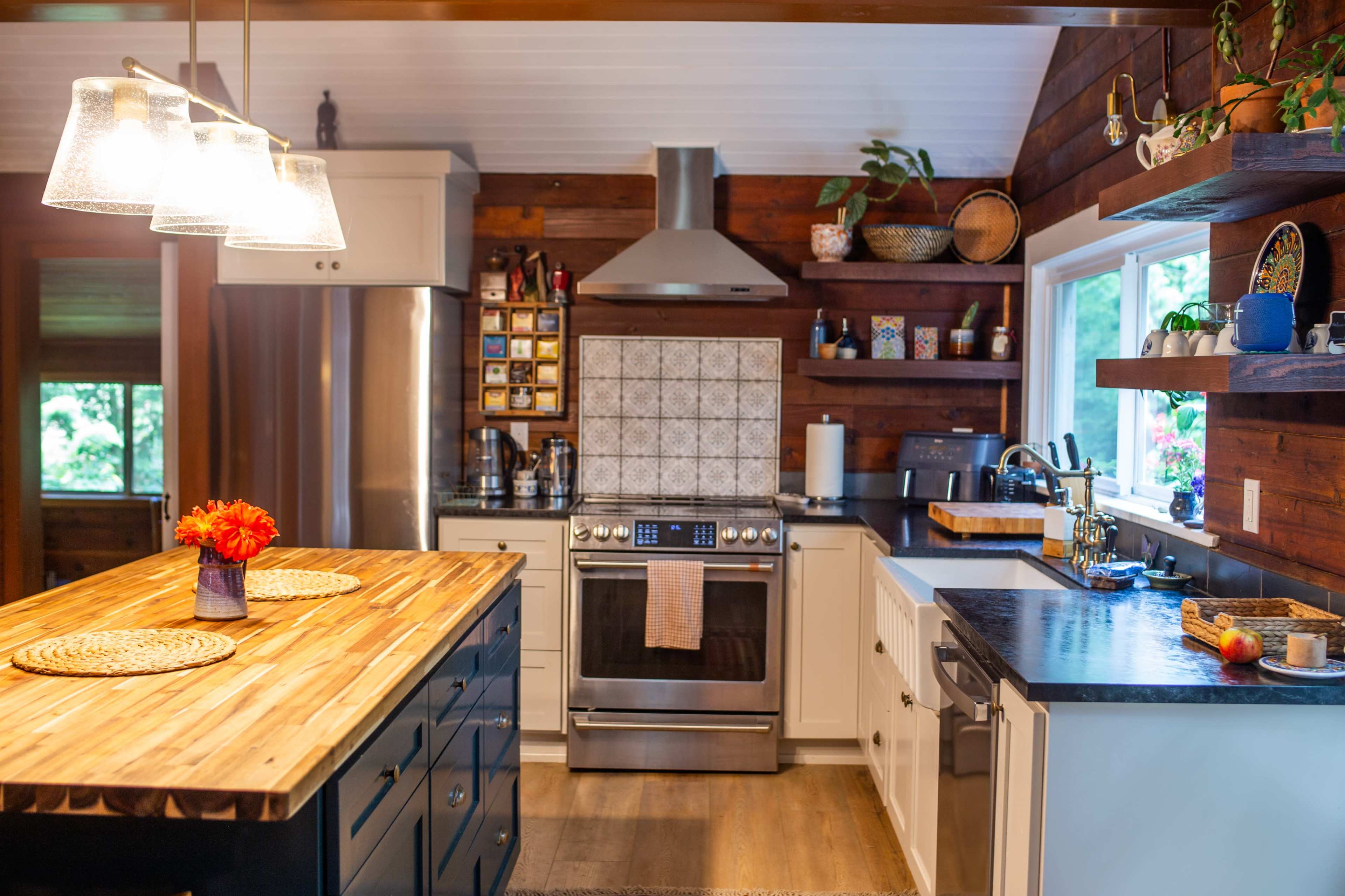 The image shows a modern kitchen featuring a wooden island, stainless steel appliances, and a mix of dark and light cabinetry with natural light filtering through the windows.
