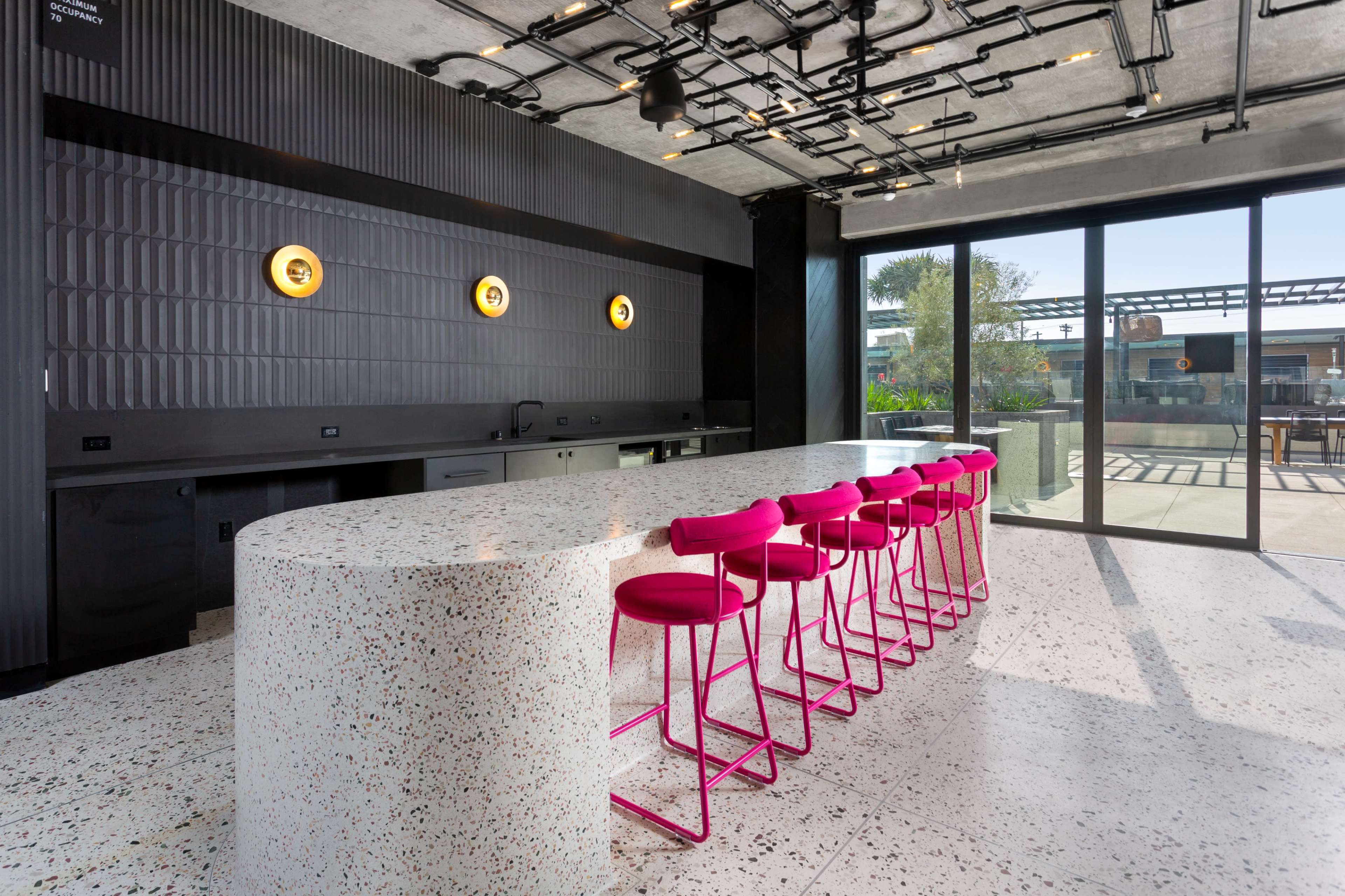 The image features a modern kitchen with a curved, speckled countertop and bright pink bar stools arranged on one side.