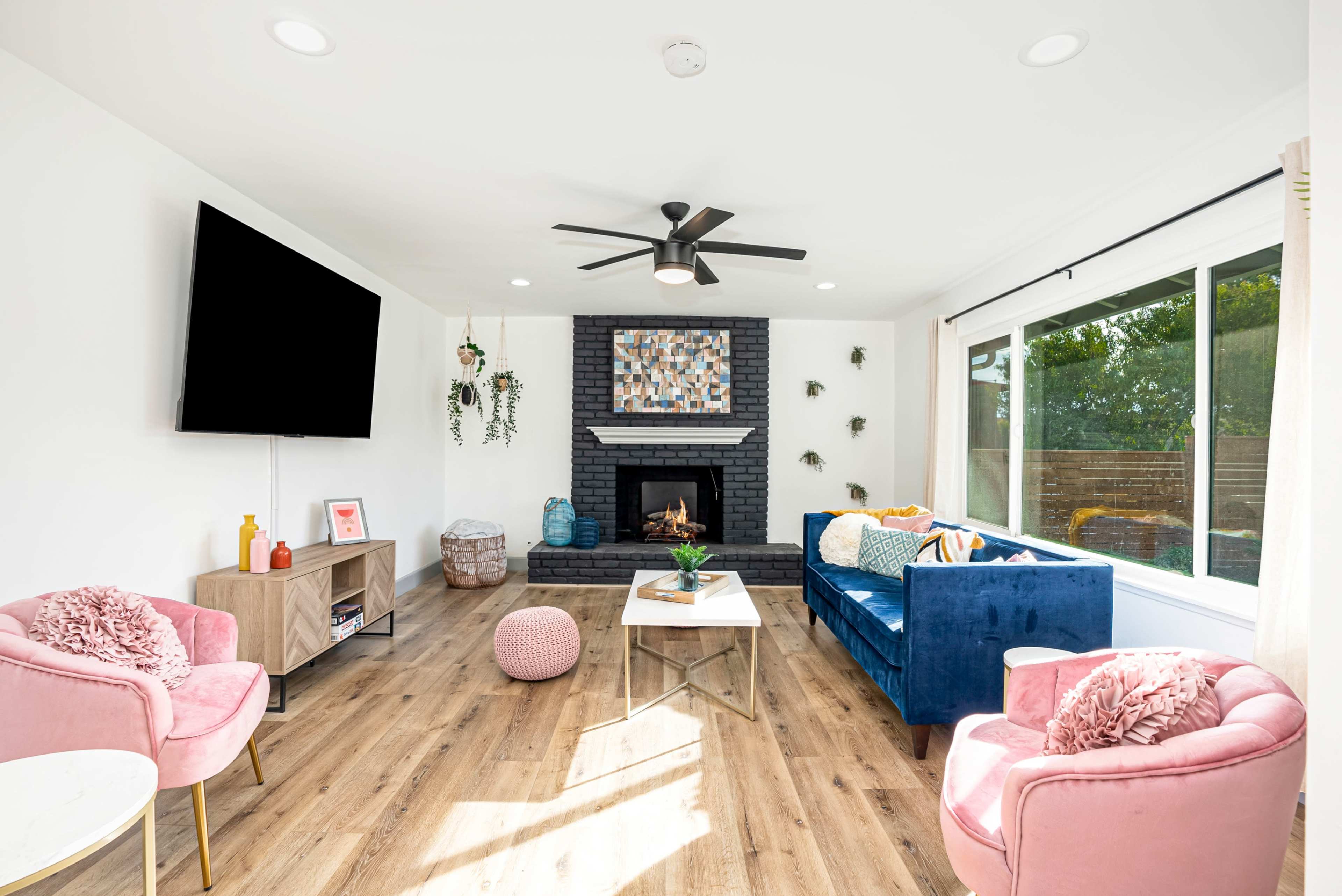 A modern living room features a blue sofa, pink armchairs, a fireplace with a black stone surround, a wall-mounted TV, and large windows allowing natural light to enter.