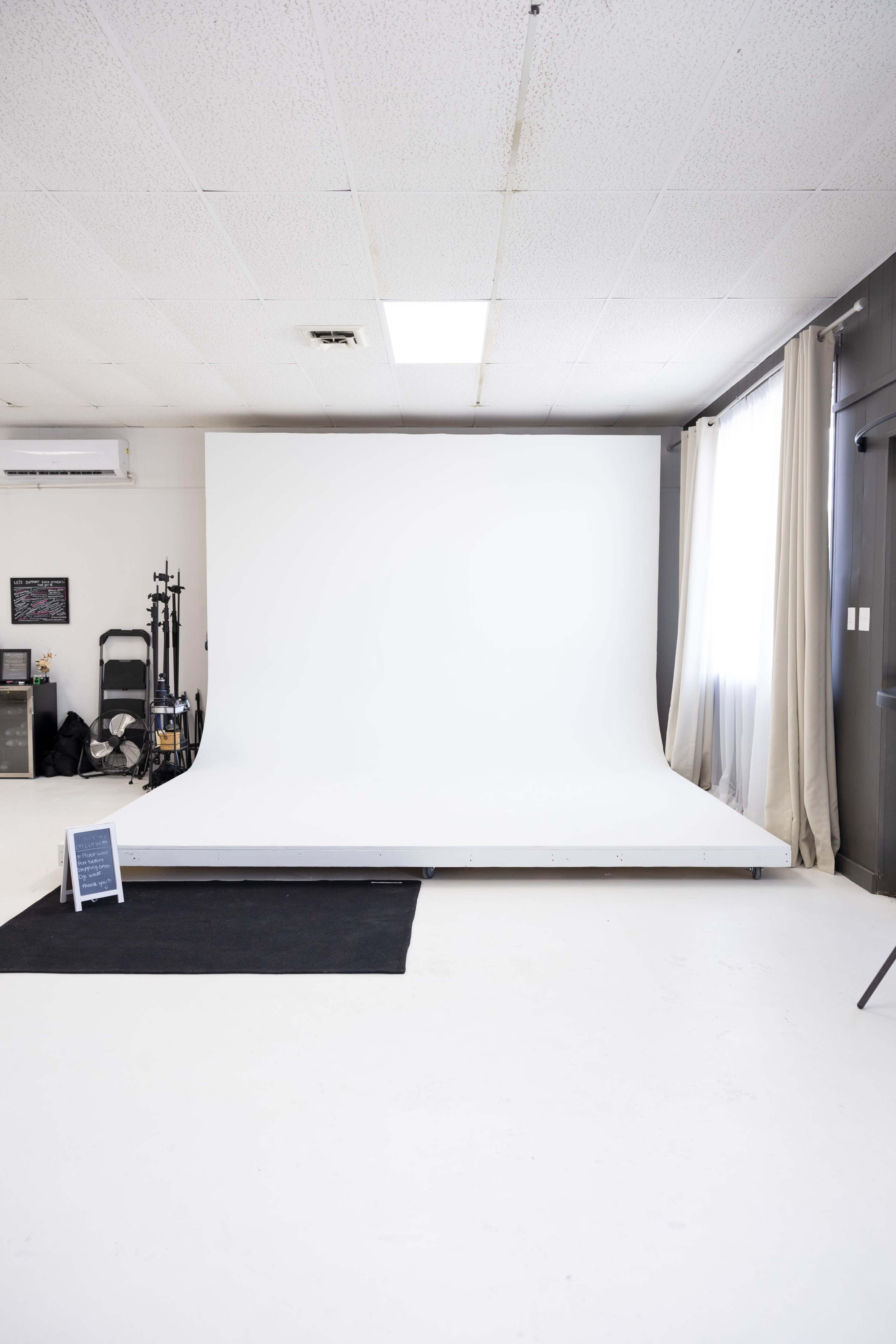 The image shows a photography studio with a blank white backdrop, a black rug, and natural light coming through a window.