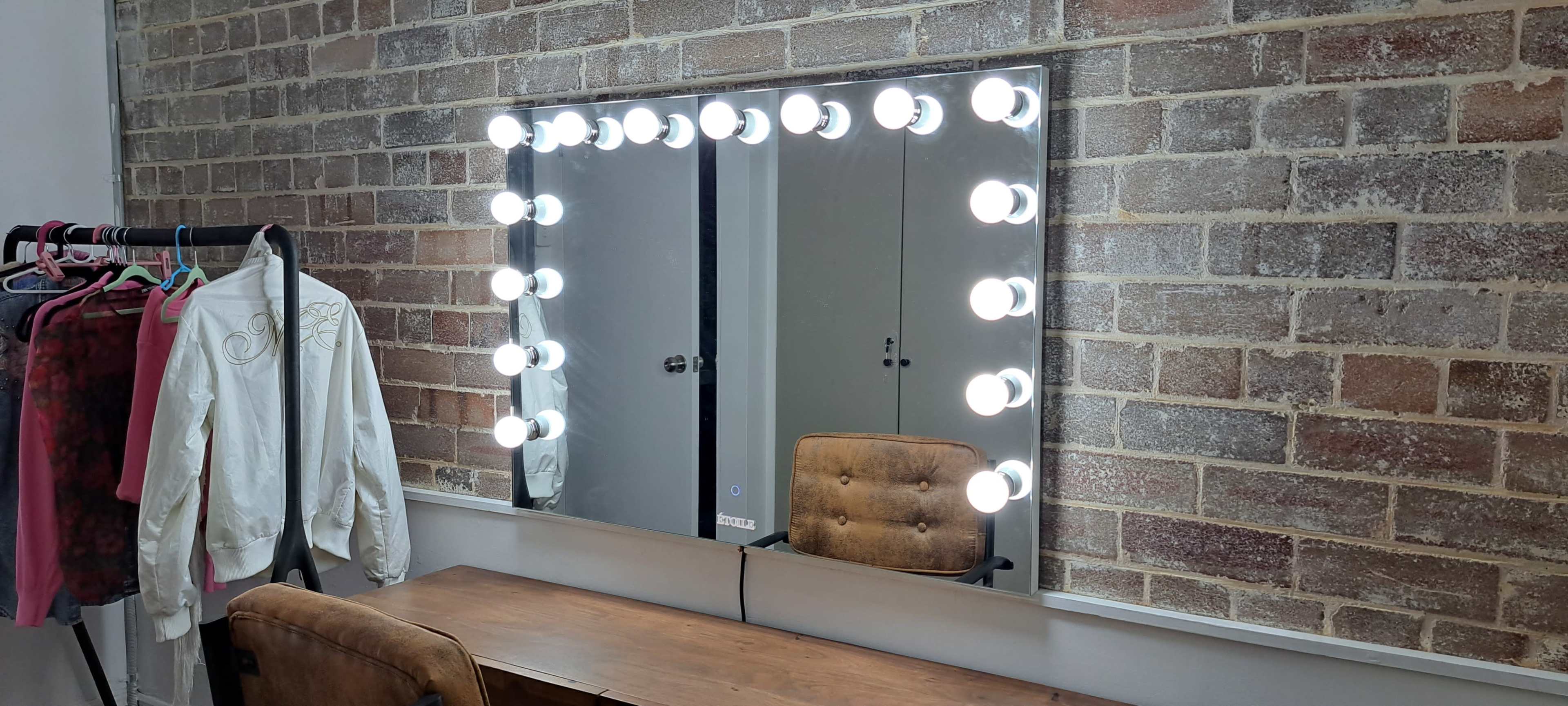 A large mirror framed by bright bulbs is mounted on a wall of exposed brick, with a wooden vanity and an upholstered chair in front, and clothing hung on a rack nearby.