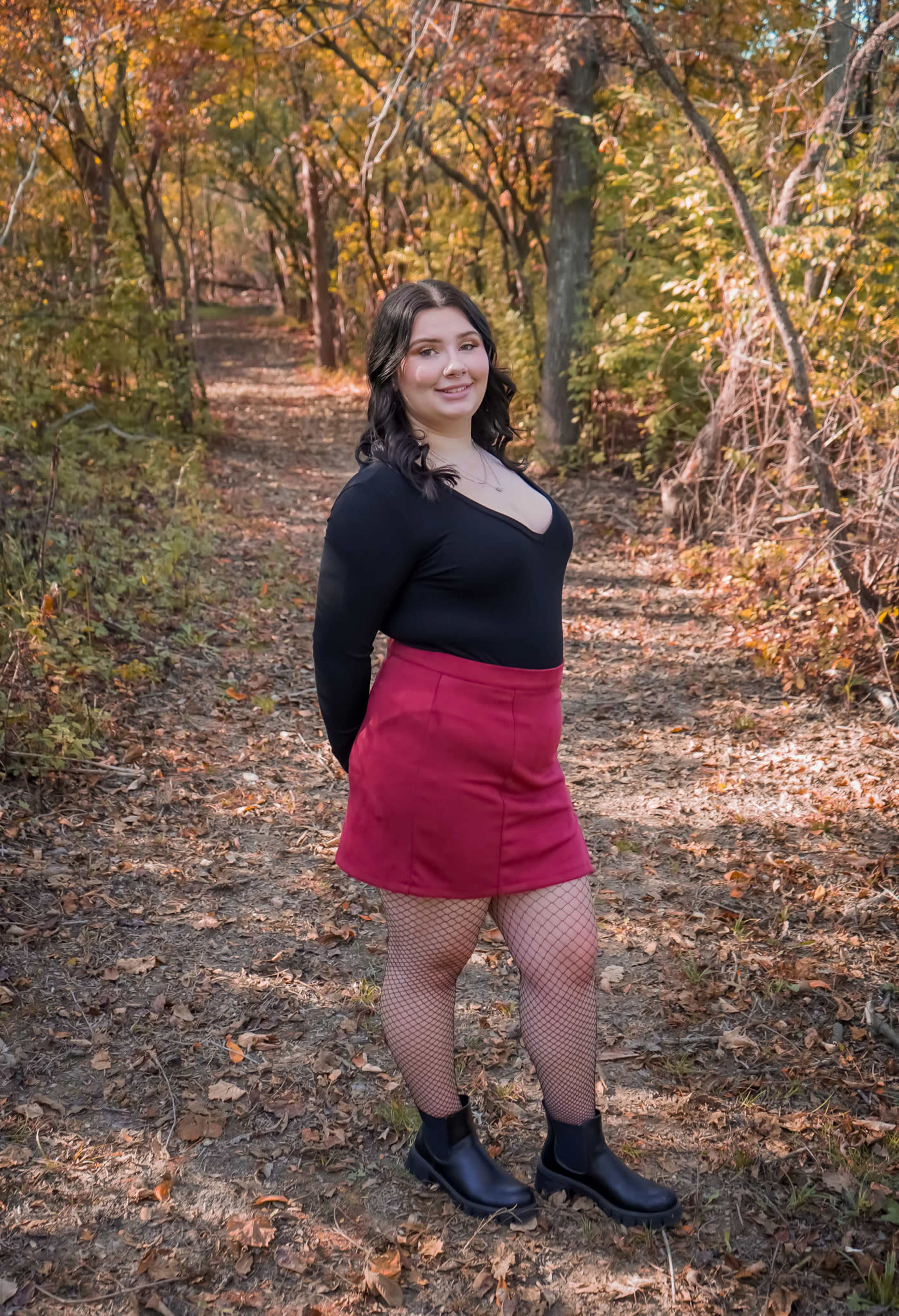 A person stands on a wooded path surrounded by autumn foliage, wearing a black top, a red skirt, and fishnet stockings.