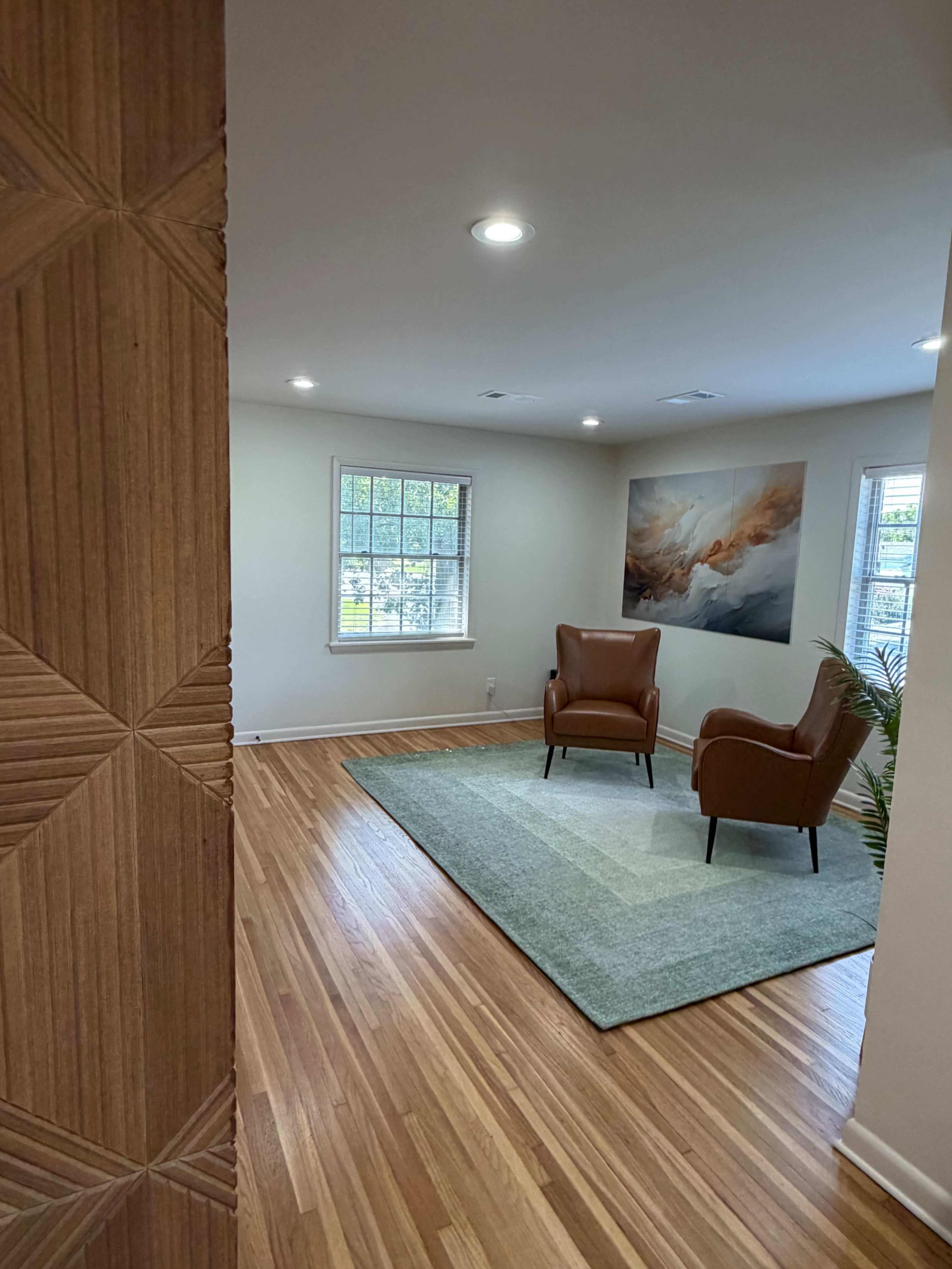 A bright room features two leather chairs on a textured rug, with a large piece of abstract art on the wall and natural light coming through the windows.