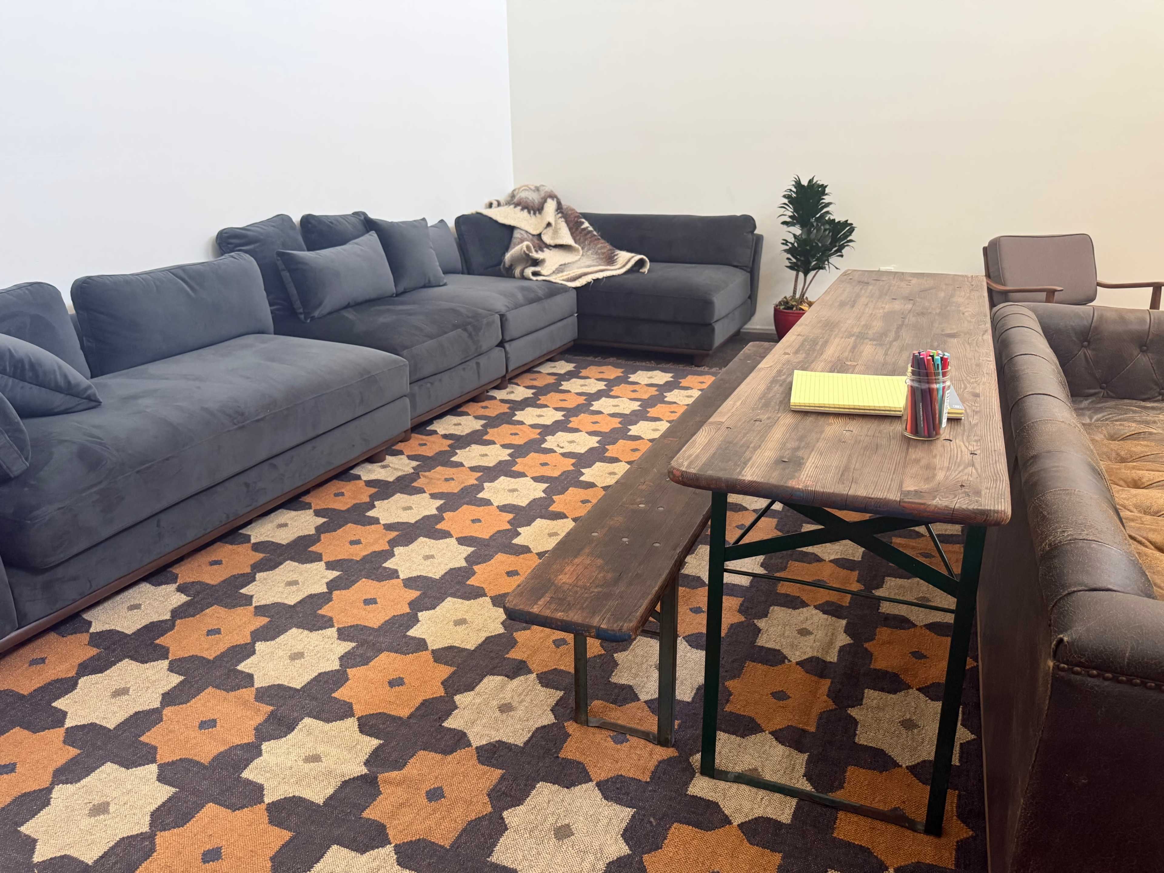 The image shows a cozy lounge area with a large gray sectional sofa, a wooden table with colorful pens, and a patterned rug.