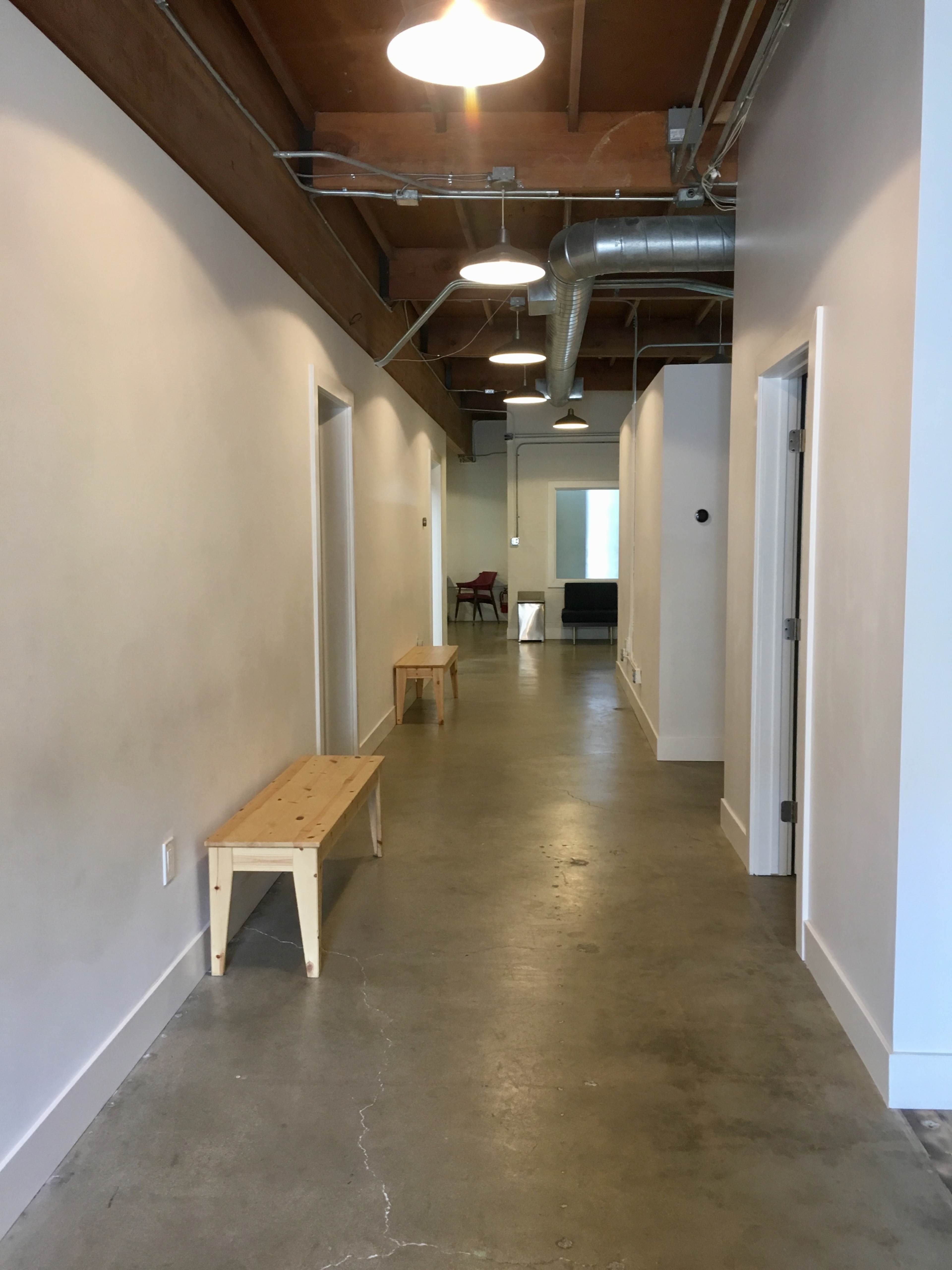 The image shows a narrow hallway with white walls, concrete flooring, two wooden benches, and several doorways on each side.