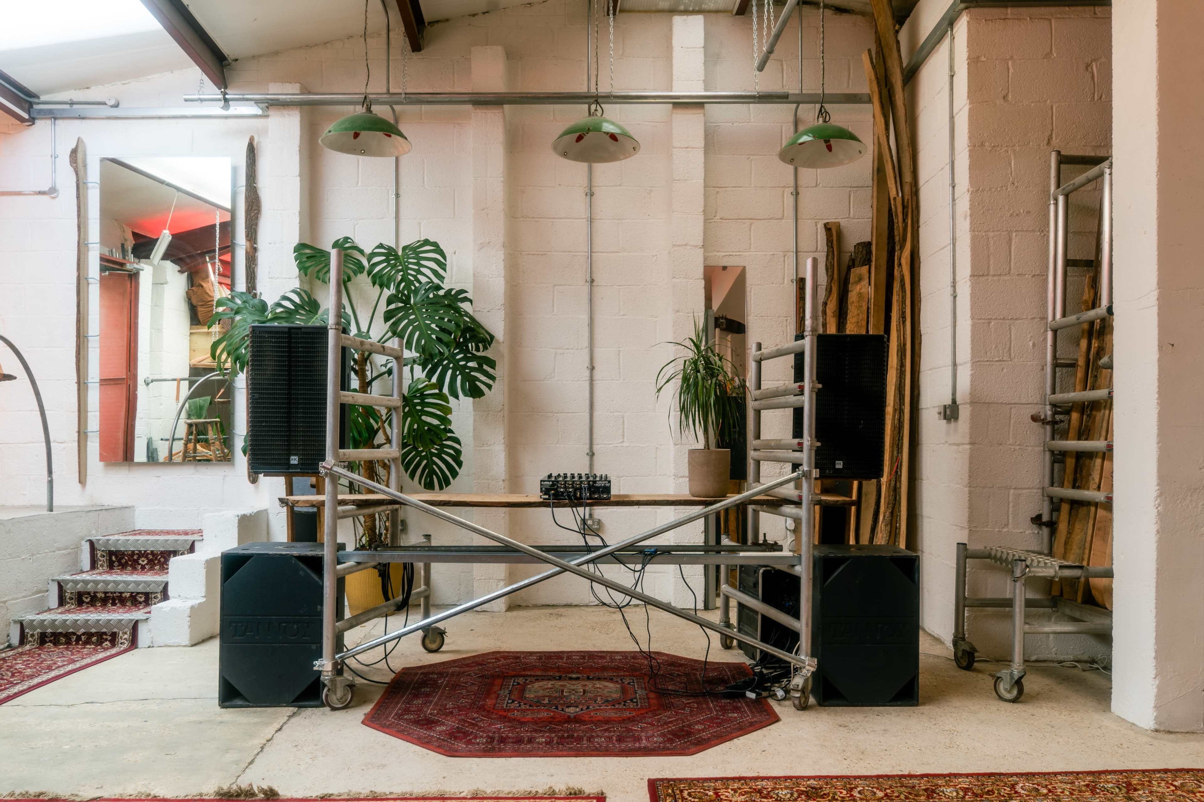 The image shows a sound setup with two large speakers on either side of a wooden table, surrounded by plants and industrial-style decor in a spacious room.