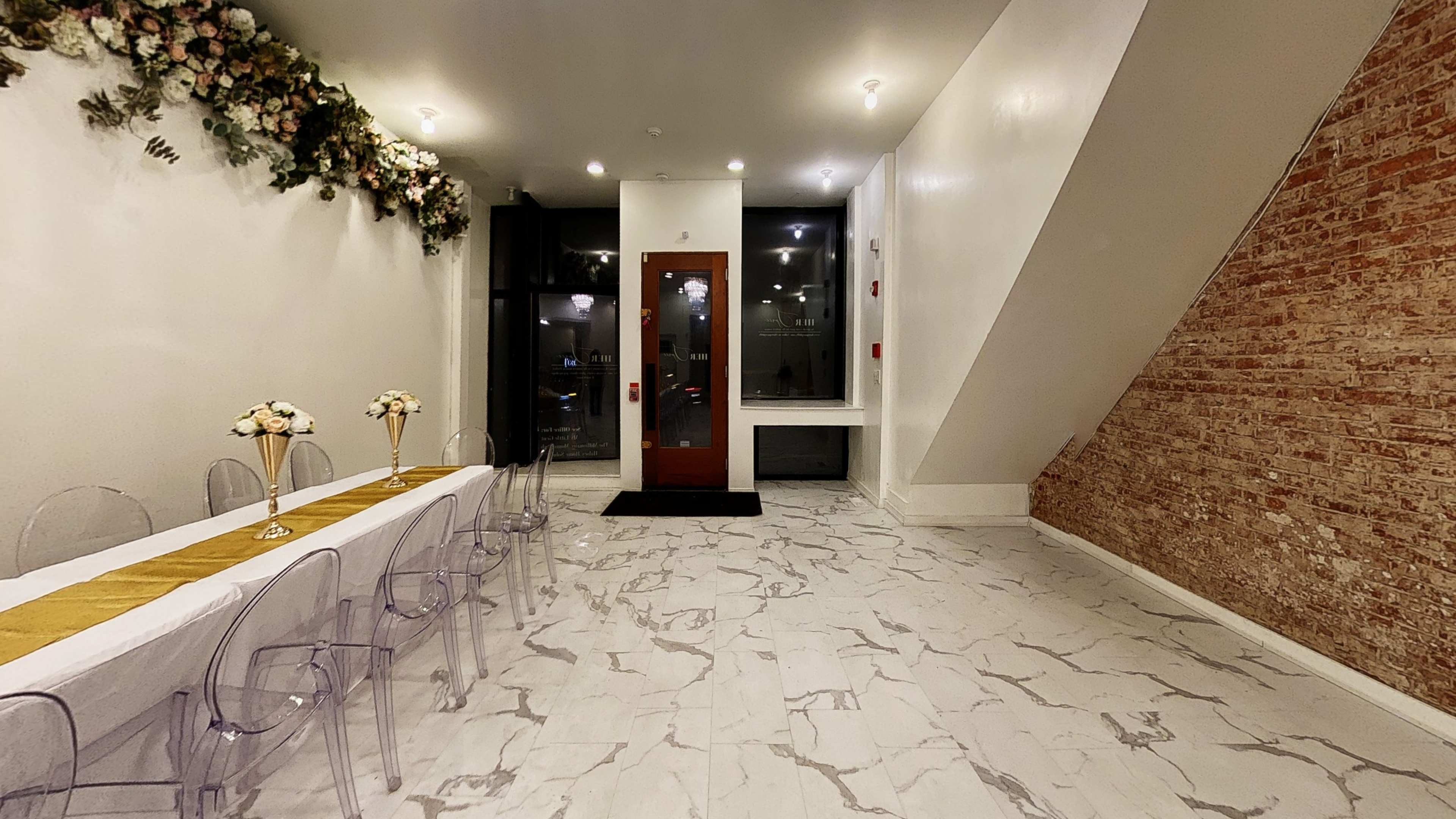 A spacious interior features a long table with clear chairs, a flower arrangement, brick walls, and an entrance at one end.
