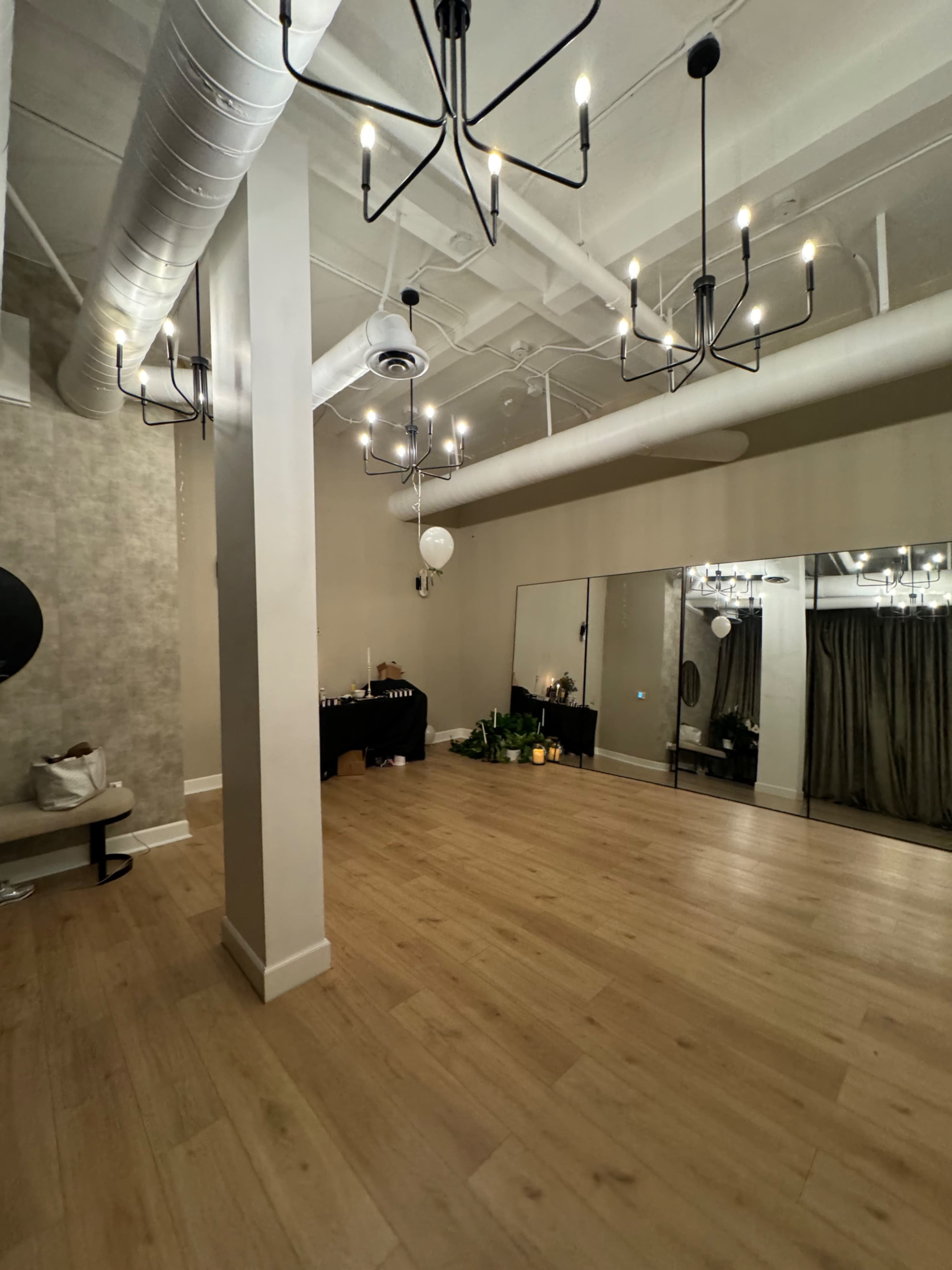 The image shows a spacious, well-lit room featuring wooden floors, multiple hanging light fixtures, a mirrored wall, and a decorative black table set against the back wall.