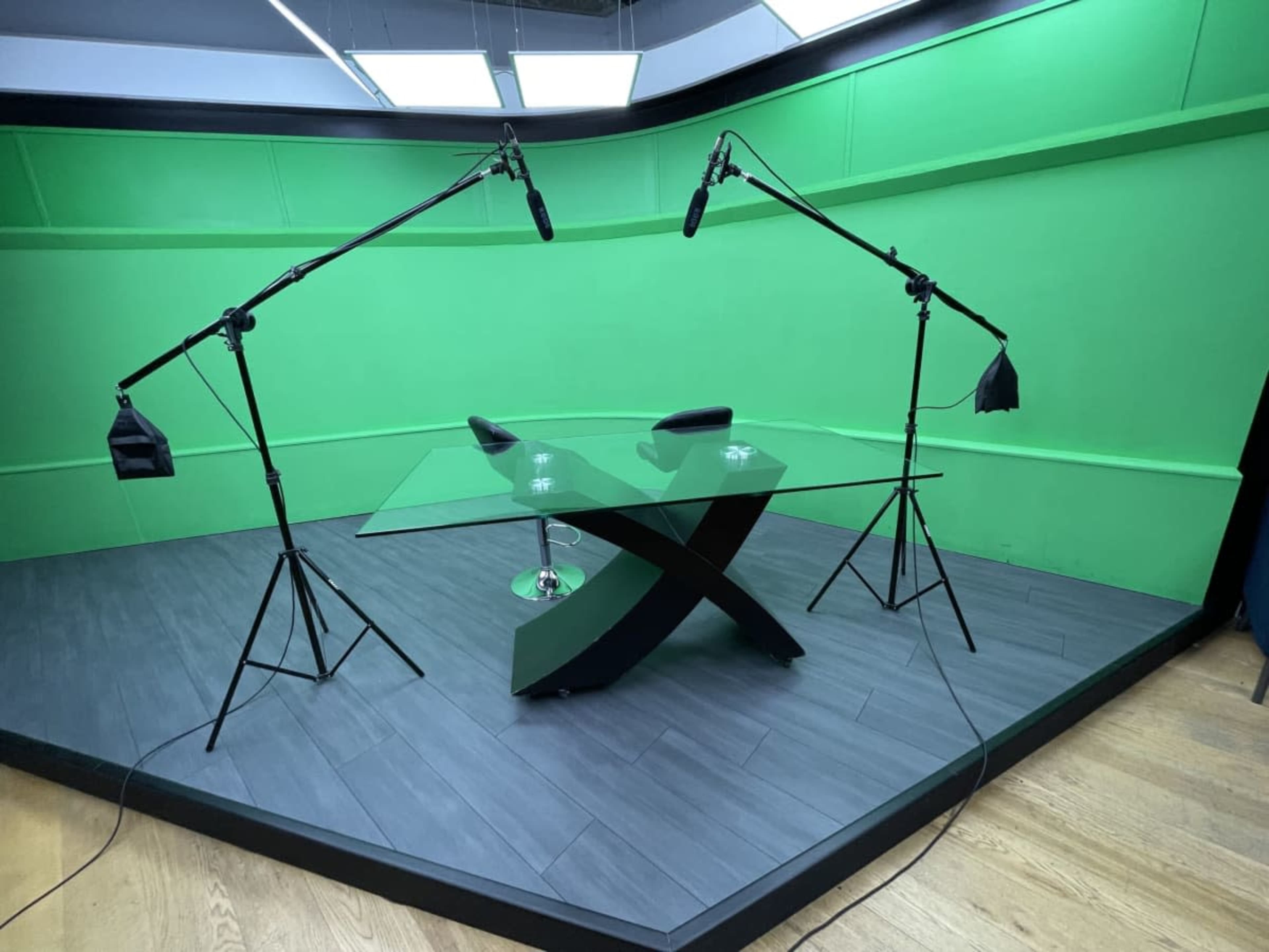 The image shows a studio setup with a glass table and two microphone boom stands positioned on either side, against a green screen backdrop.