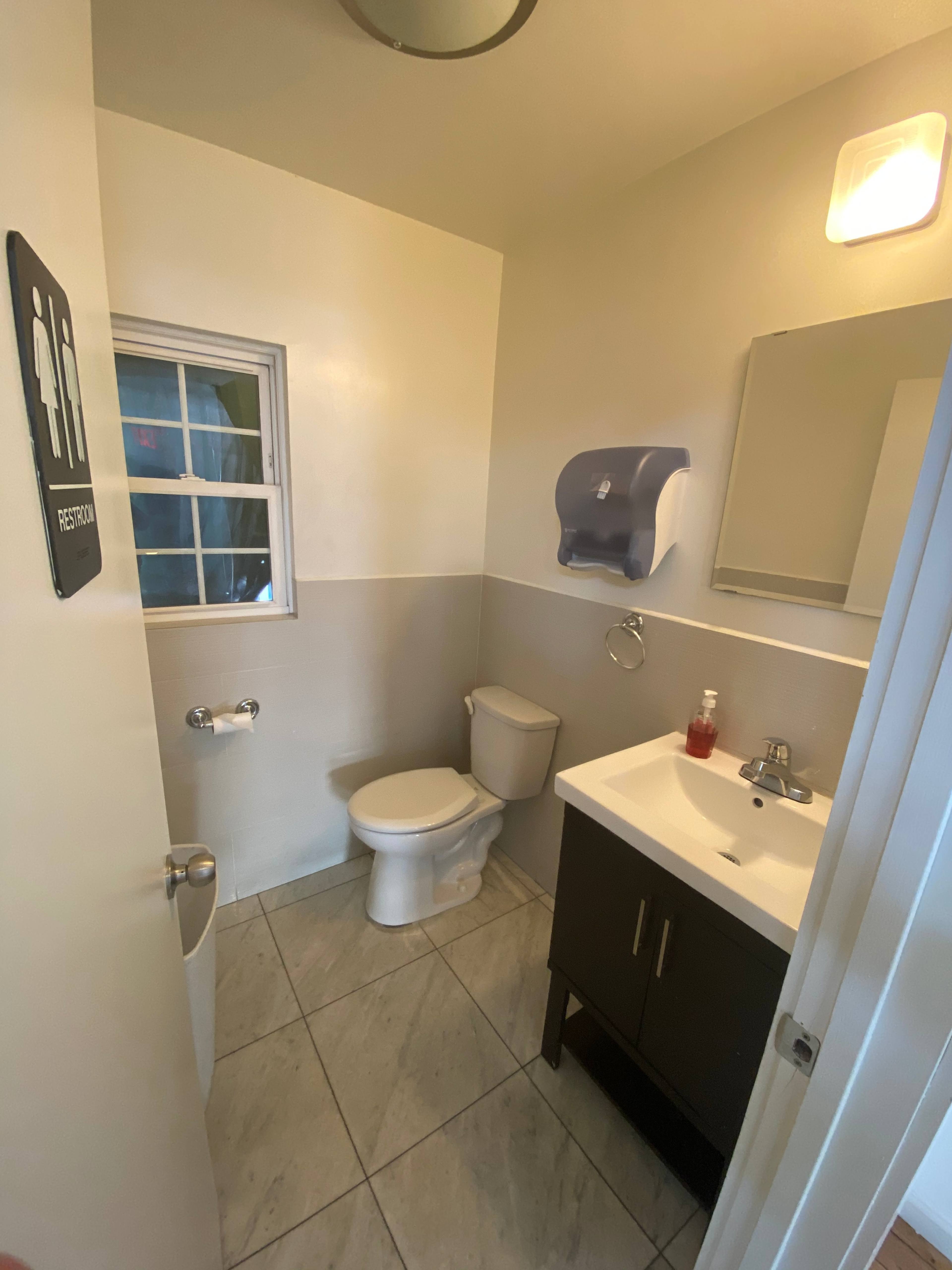 A small restroom featuring a toilet, sink with a cabinet, a mirror, and a window.