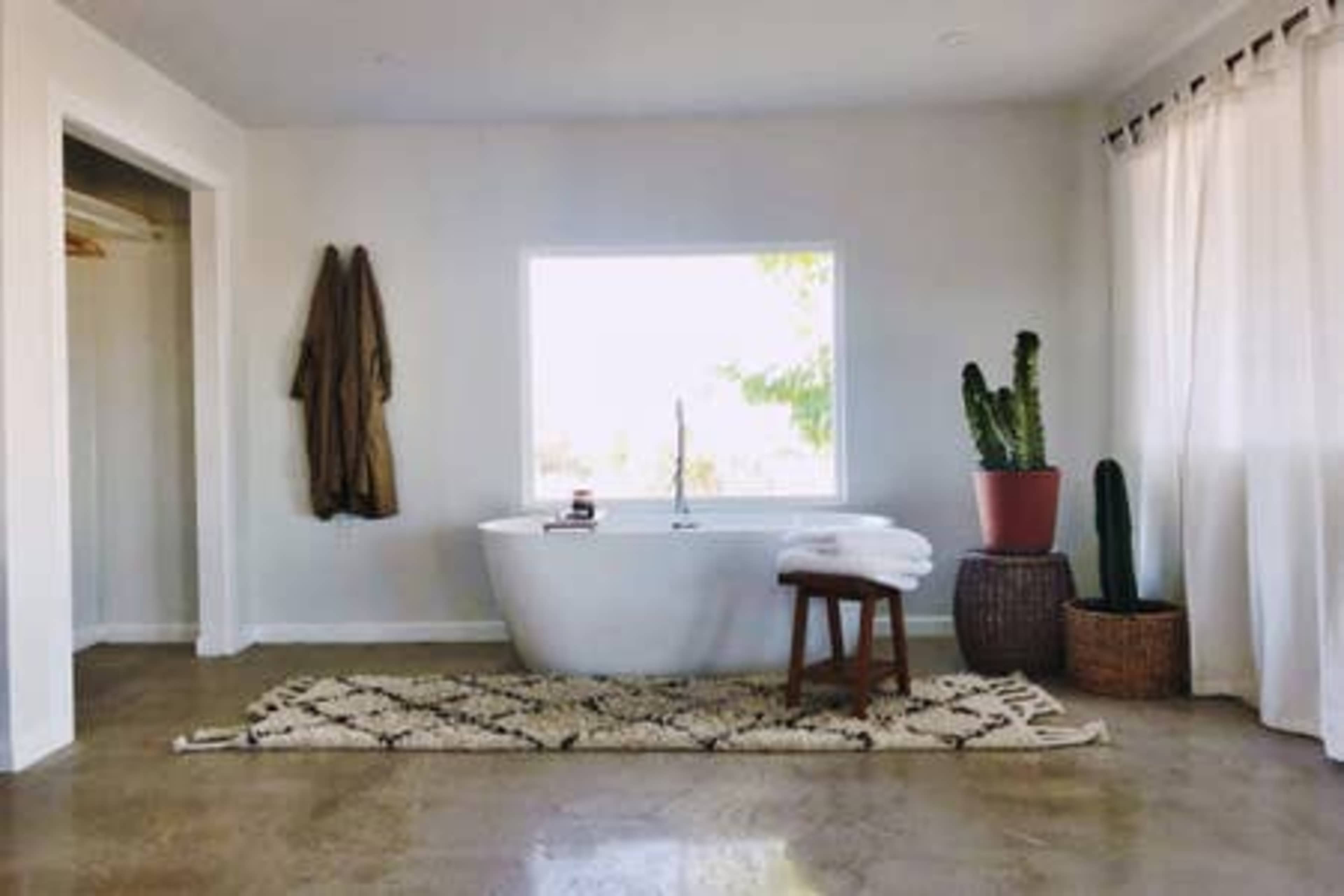 A bright bathroom featuring a freestanding tub, a potted cactus, a stool, and a large window allowing natural light to enter.