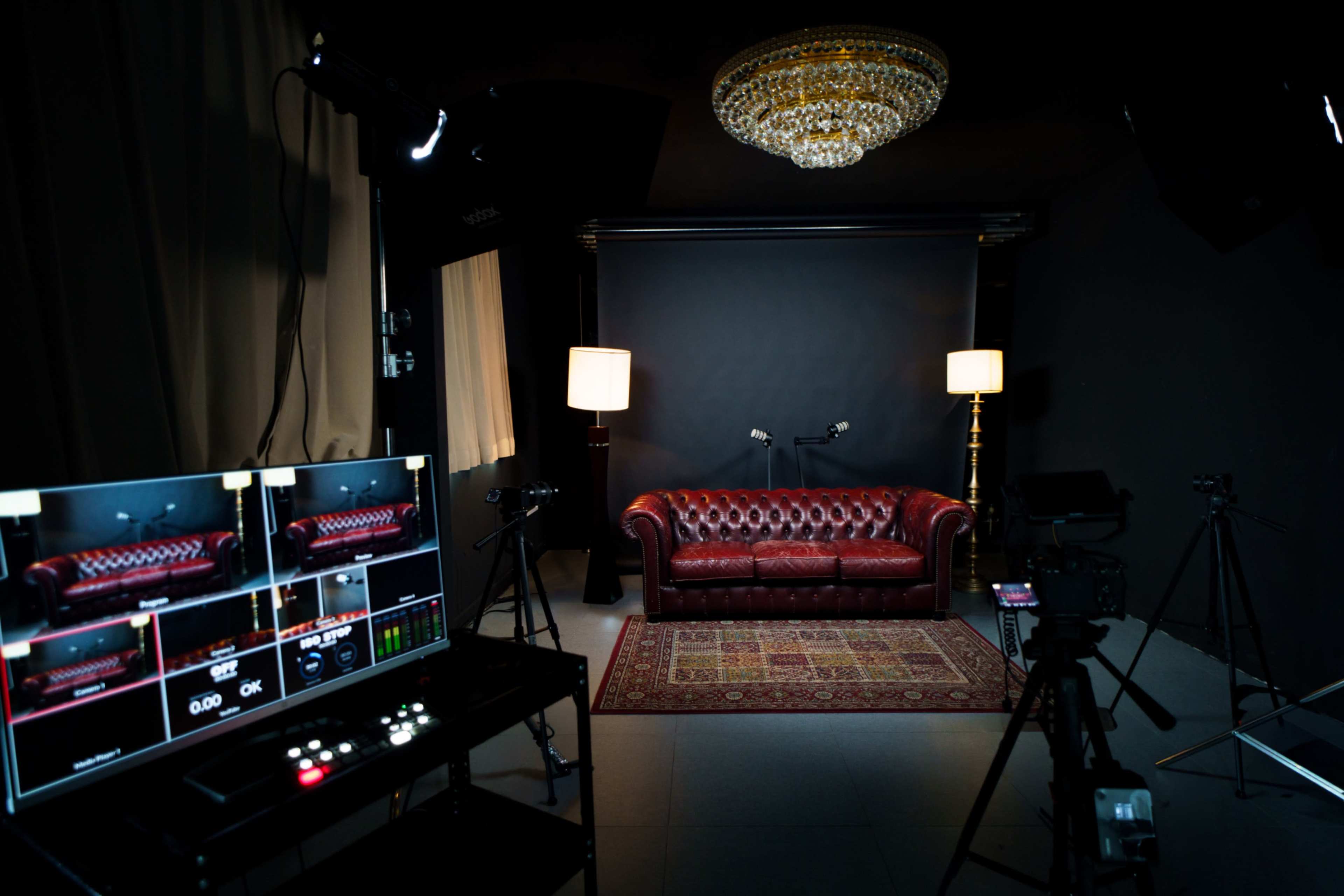 The image shows a dimly lit recording studio with a leather couch, decorative rug, and multiple cameras set up, accompanied by lighting equipment.