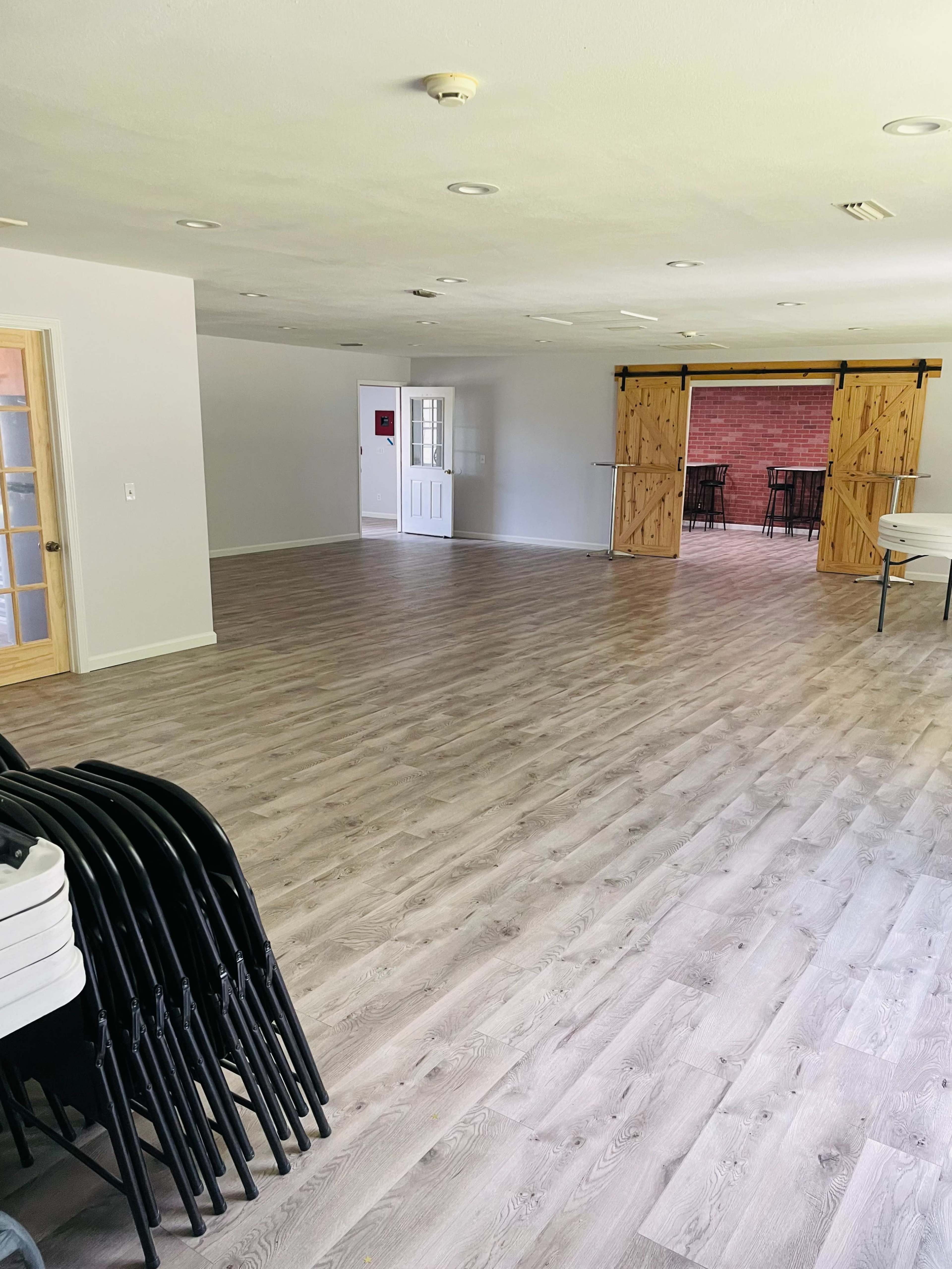 A spacious interior room with light wooden flooring, empty walls, and a set of sliding barn doors leading to a brick patio area.