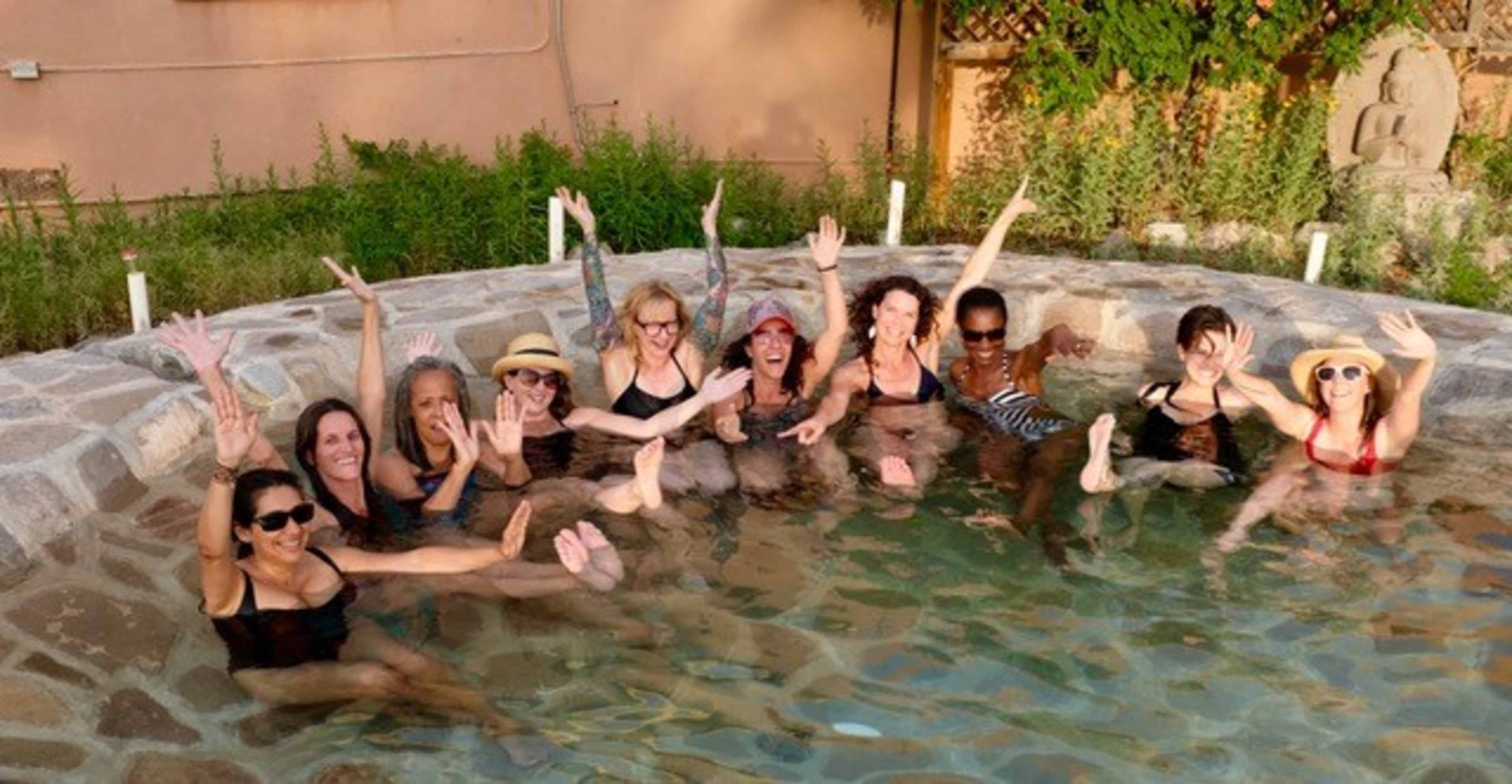 A group of women is sitting in a stone pool, smiling and waving while enjoying the water.