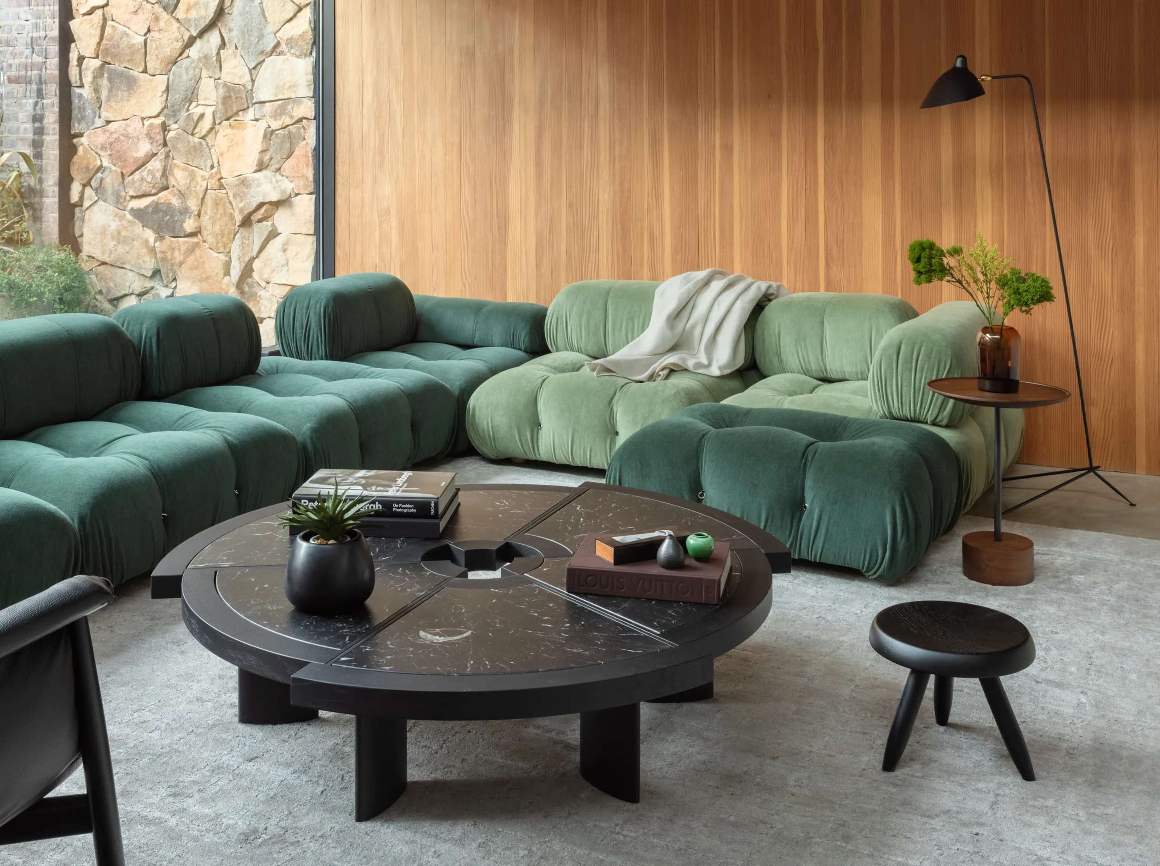 The image depicts a modern living room featuring a green, sectional sofa, a round black coffee table, and a wooden accent wall.