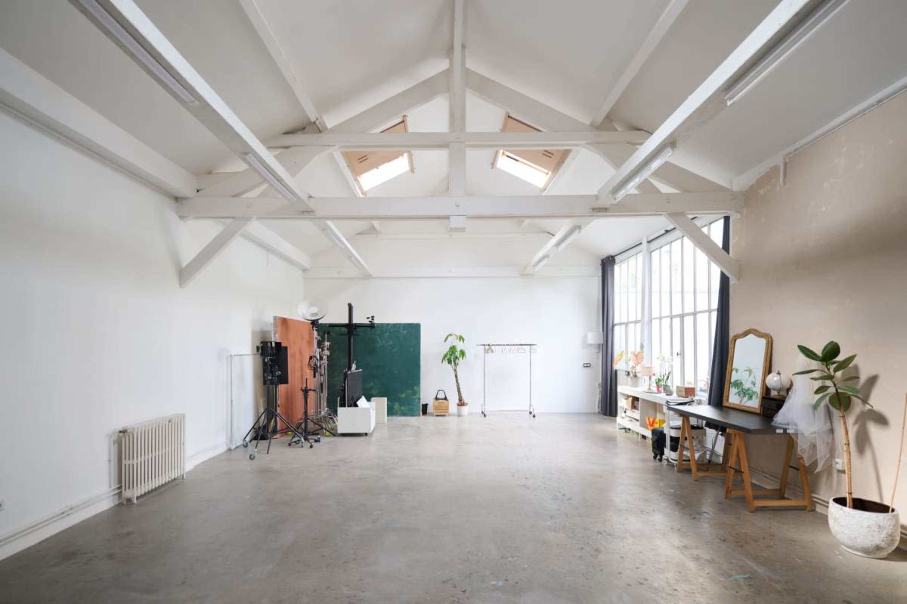 The image shows a spacious, empty room with high ceilings, large windows, and minimal furniture, including a desk and some studio equipment.