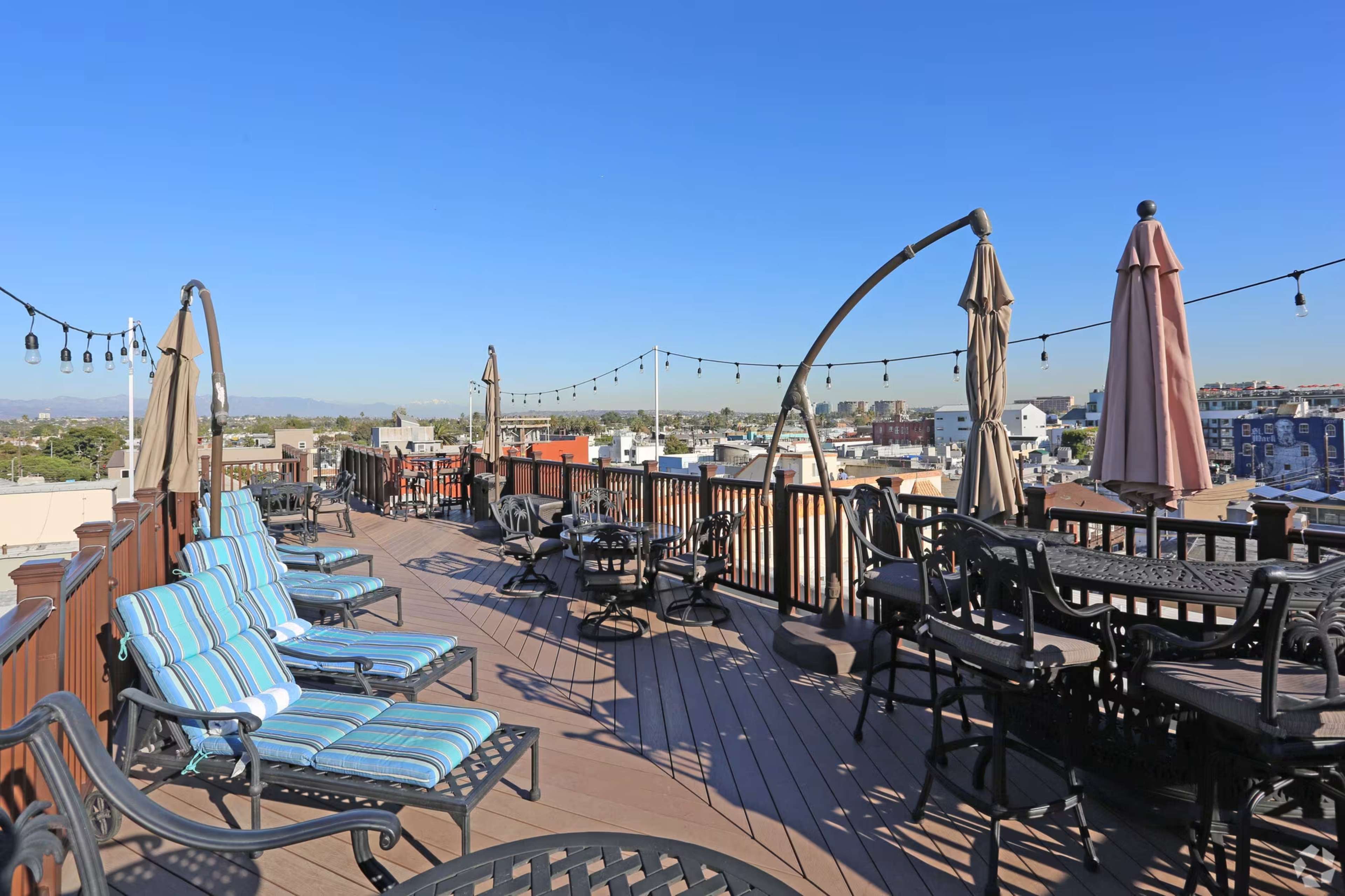 The image shows a rooftop deck with lounge chairs, tables, and large umbrellas, surrounded by a city skyline under a clear blue sky.
