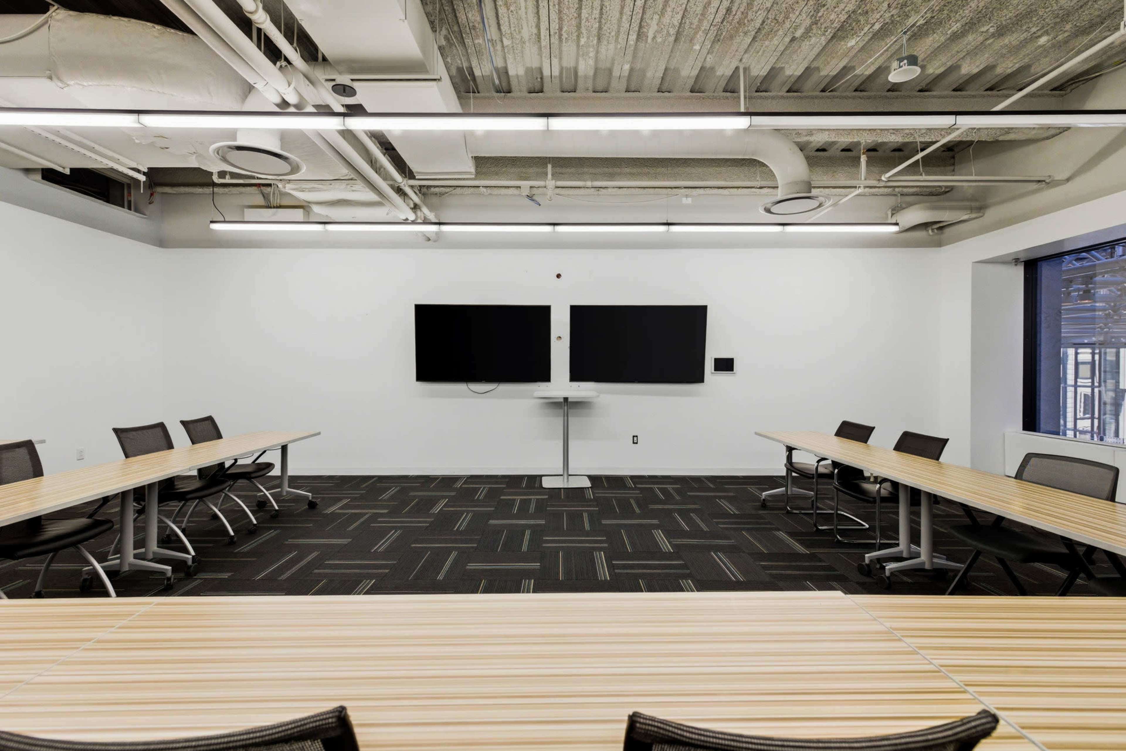 The image shows a modern conference room with two large screens on the wall and several tables arranged in a U-shape, accompanied by black chairs.