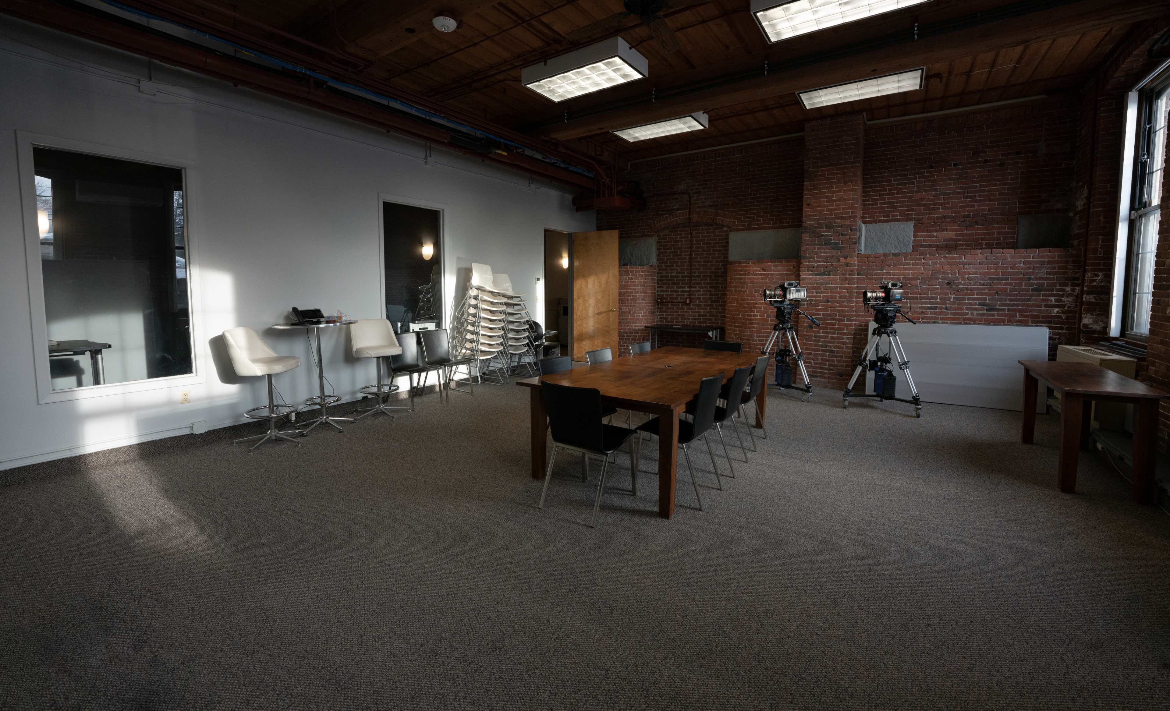 A spacious meeting room featuring a long wooden table, black chairs, and two video cameras positioned on tripods, with exposed brick walls and a carpeted floor.