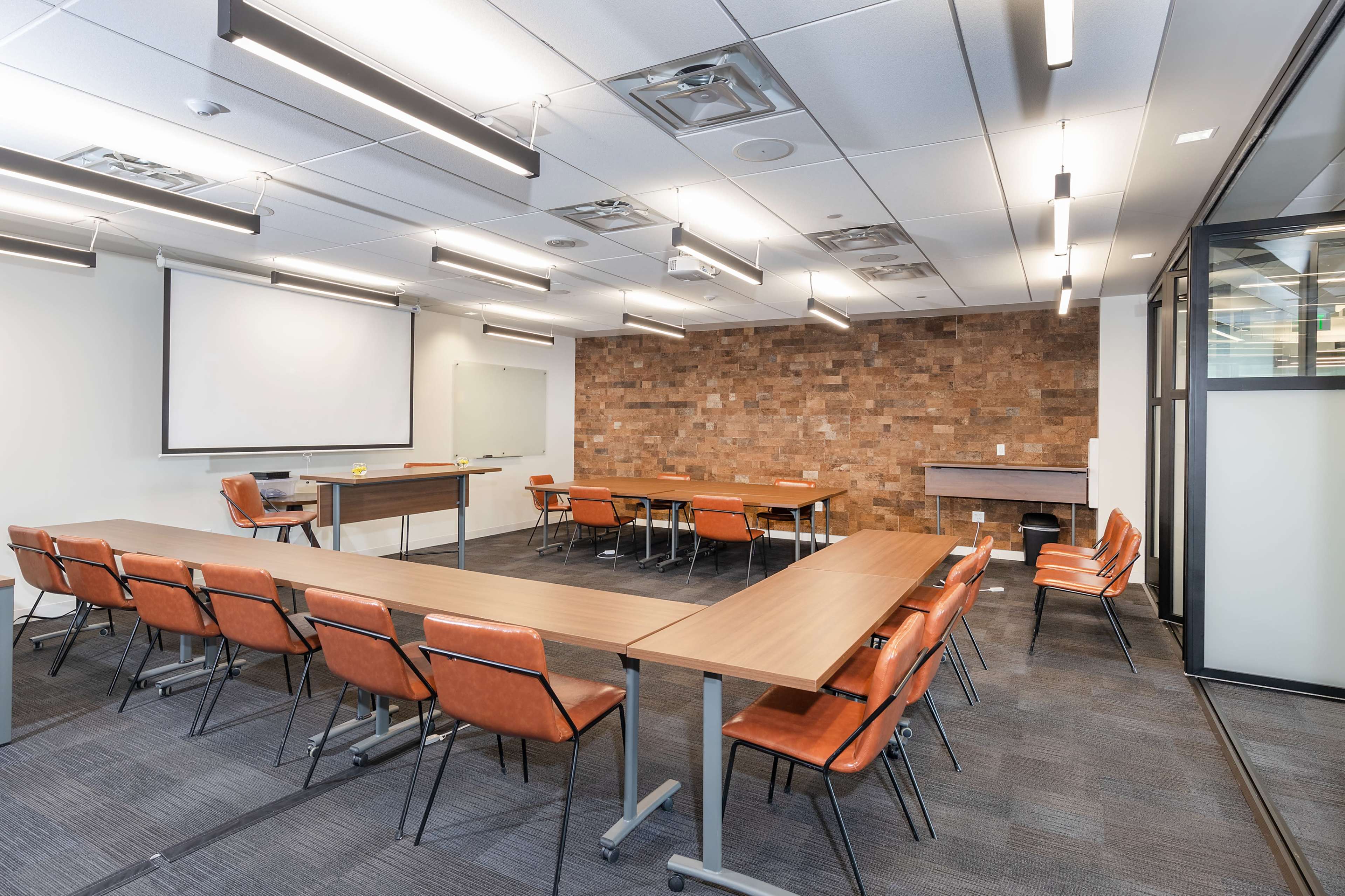 A modern conference room features long wooden tables surrounded by orange chairs, with a presentation screen at one end and a textured stone wall.