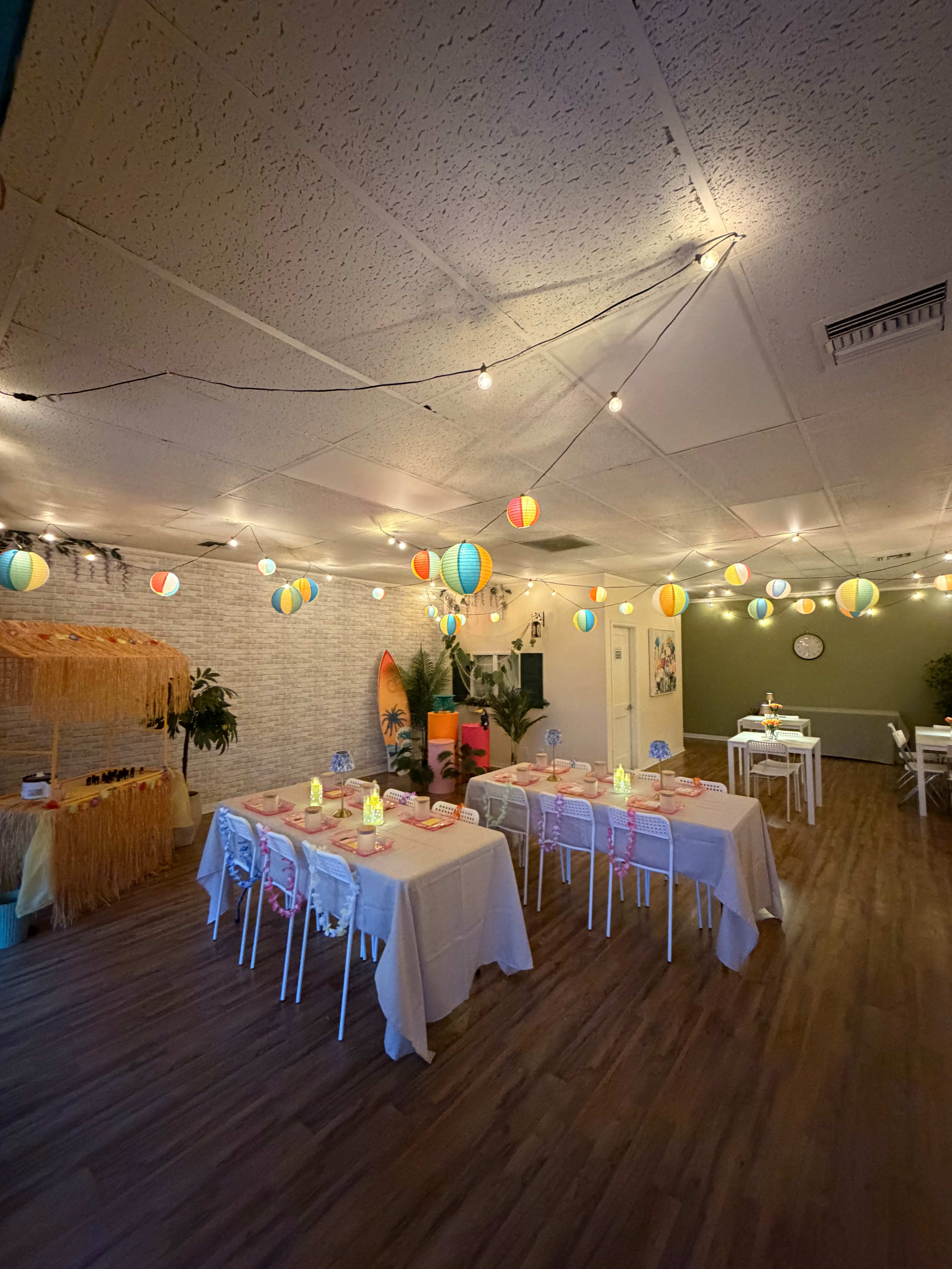 The image shows a decorated indoor party space with white tables set for dining, colorful hanging lanterns, and a surfboard in the background.