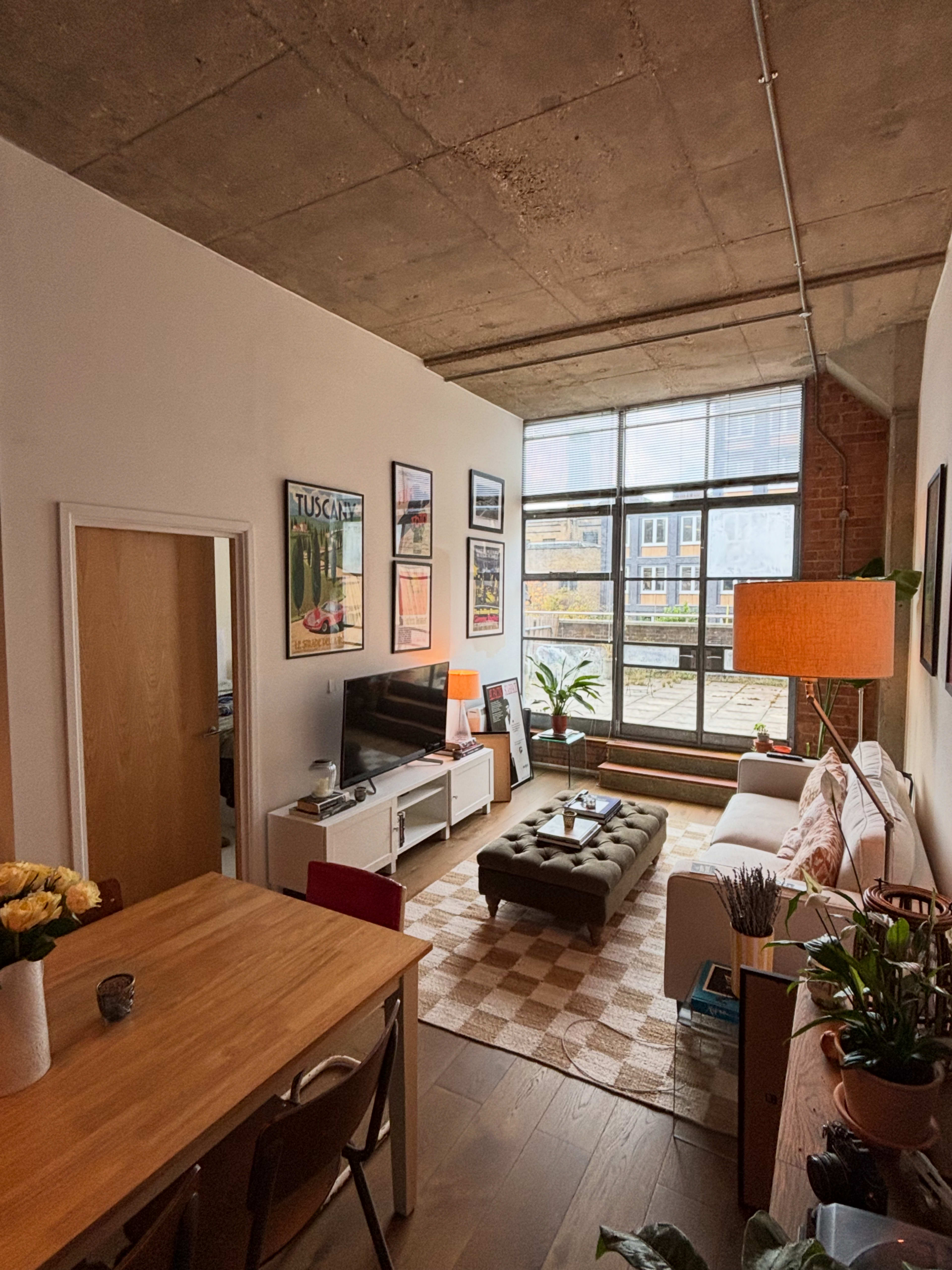 The image shows a modern apartment living area with large windows, a comfortable sofa, a dining table, and various decorative elements.
