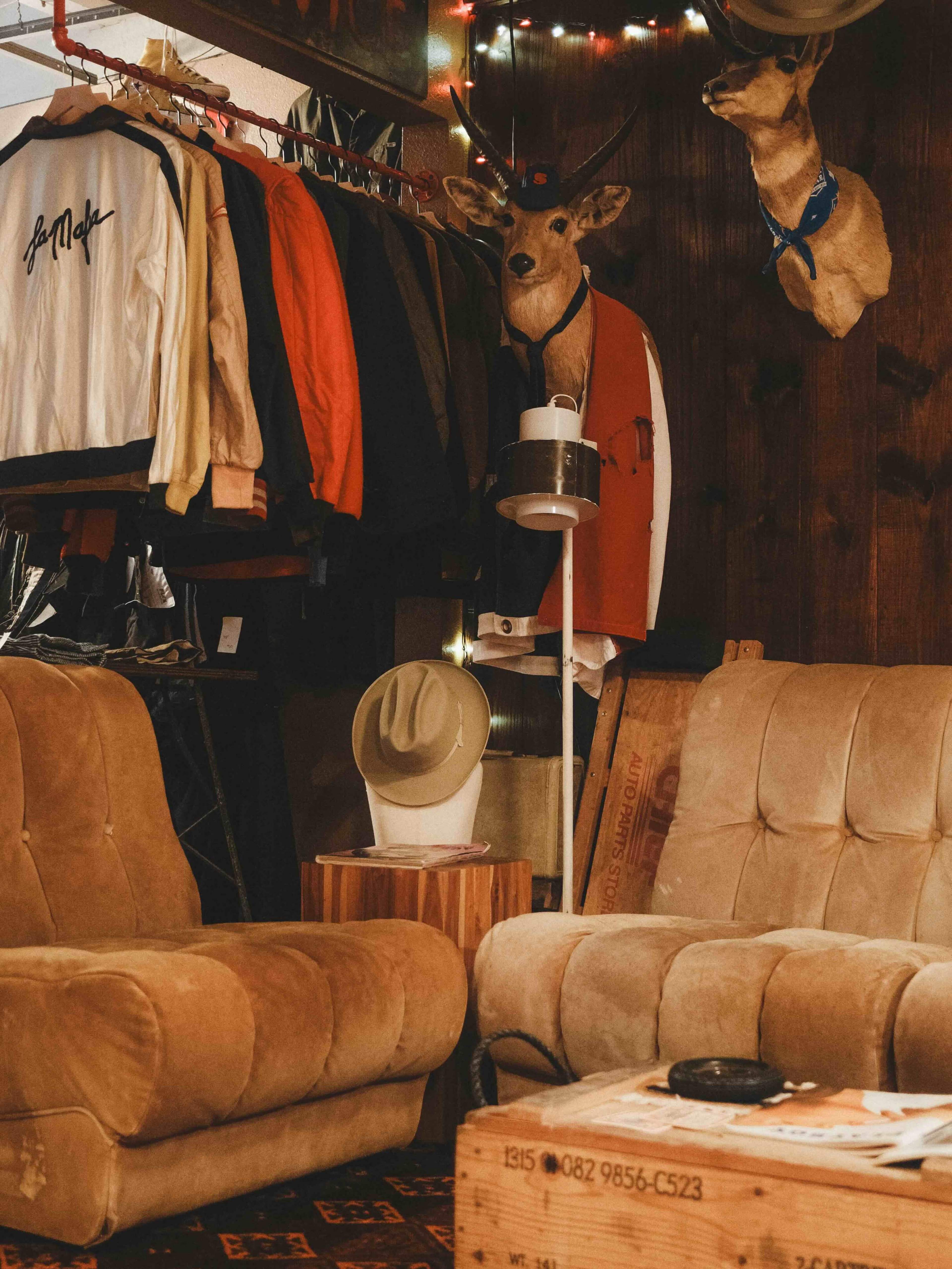 The scene features two beige couches facing each other in a room with clothing hanging on a rack, mounted deer heads on the wall, and various decorative items.
