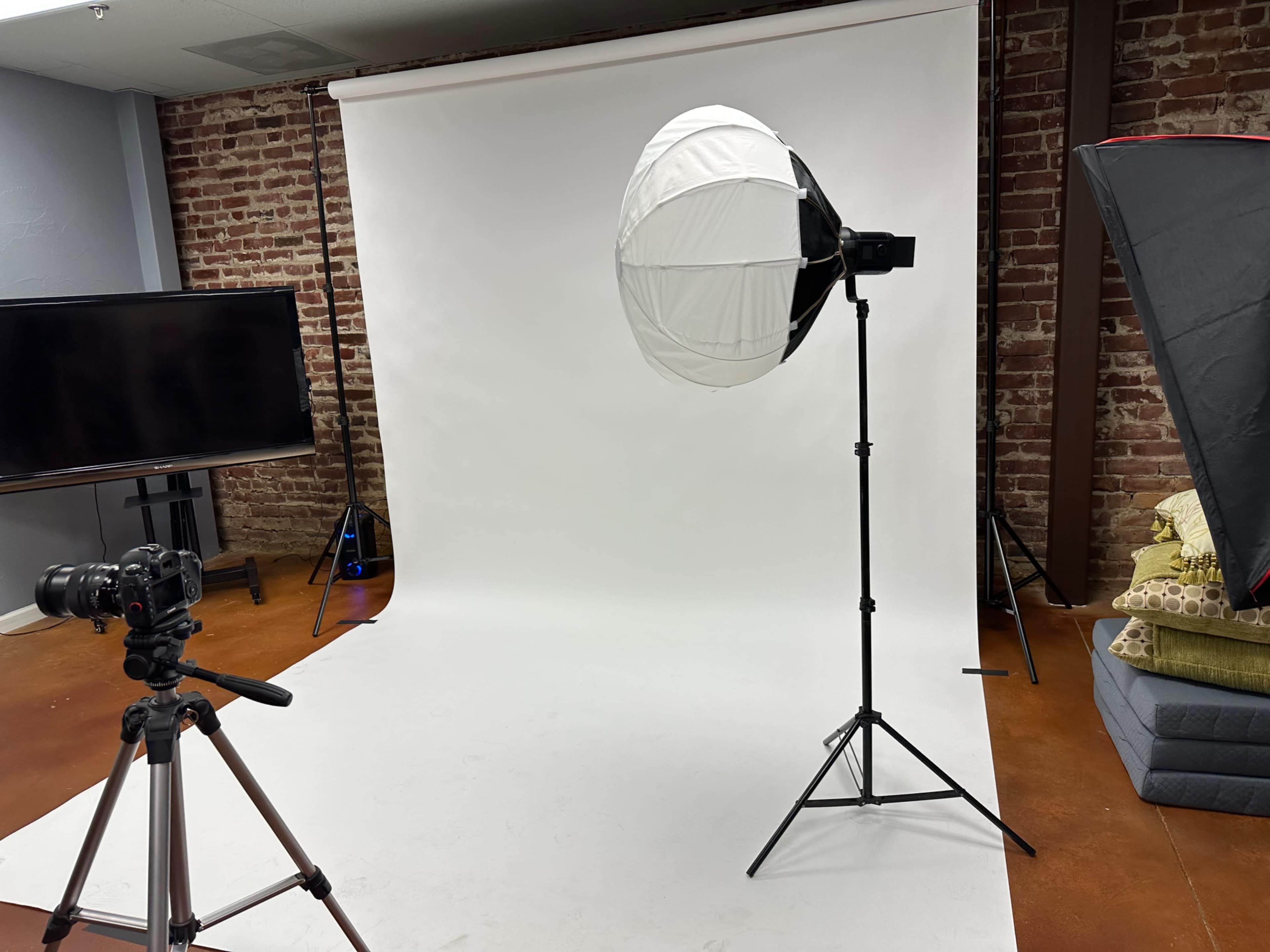 The image shows a photography studio with a camera on a tripod facing a white backdrop lit by a softbox and another light source.